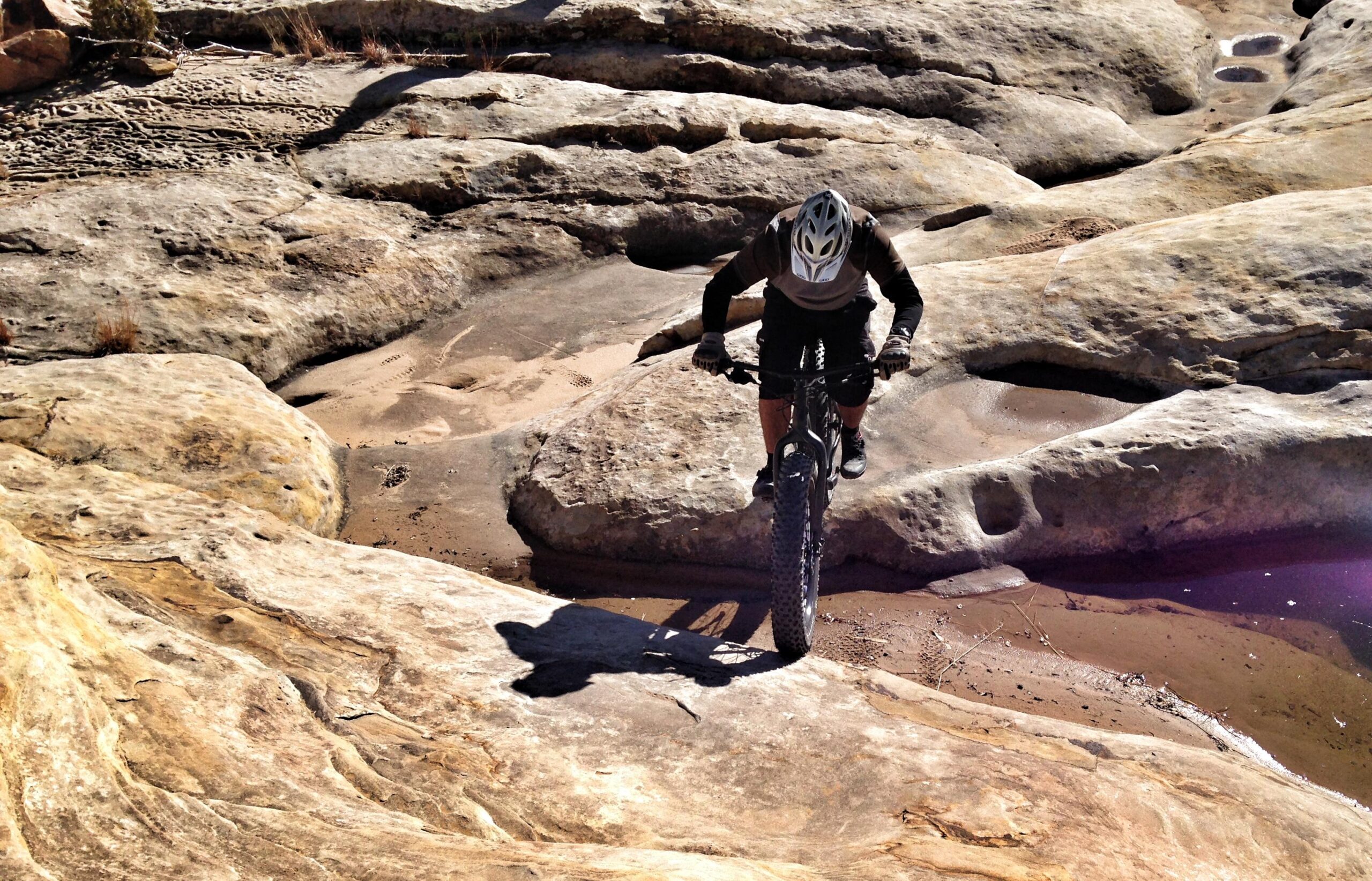 Specialized Fatboy: A mountain biker navigating a rocky terrain, featuring large, rough stones and patches of sand. The biker is leaning forward, using careful balance while riding over the rocky surface under bright sunlight.