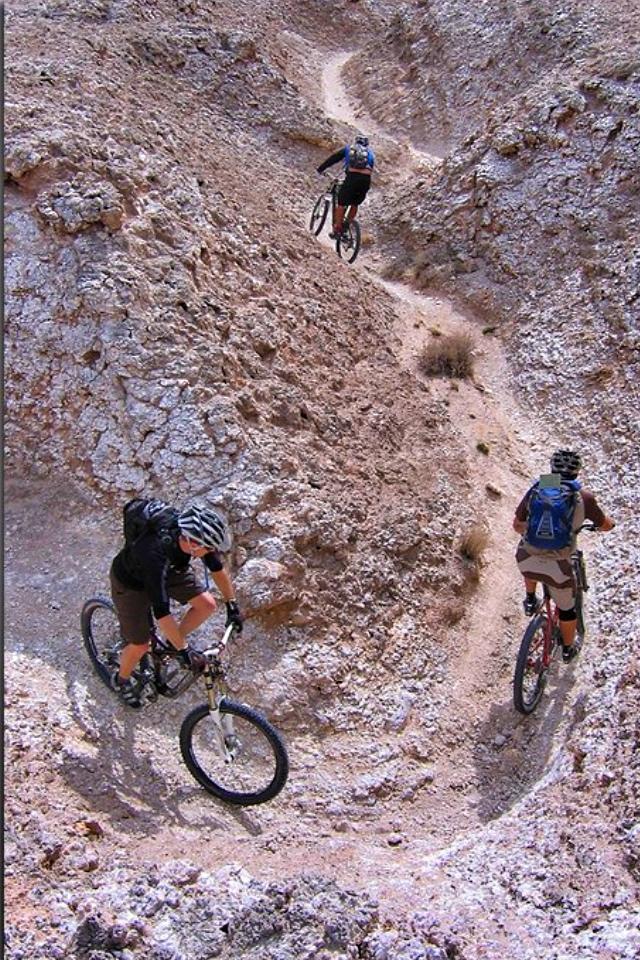 Three mountain bikers navigate a winding dirt trail through rocky terrain, showcasing their biking skills against a backdrop of rugged hills. White Mesa Bike Trails mountain bike trail.