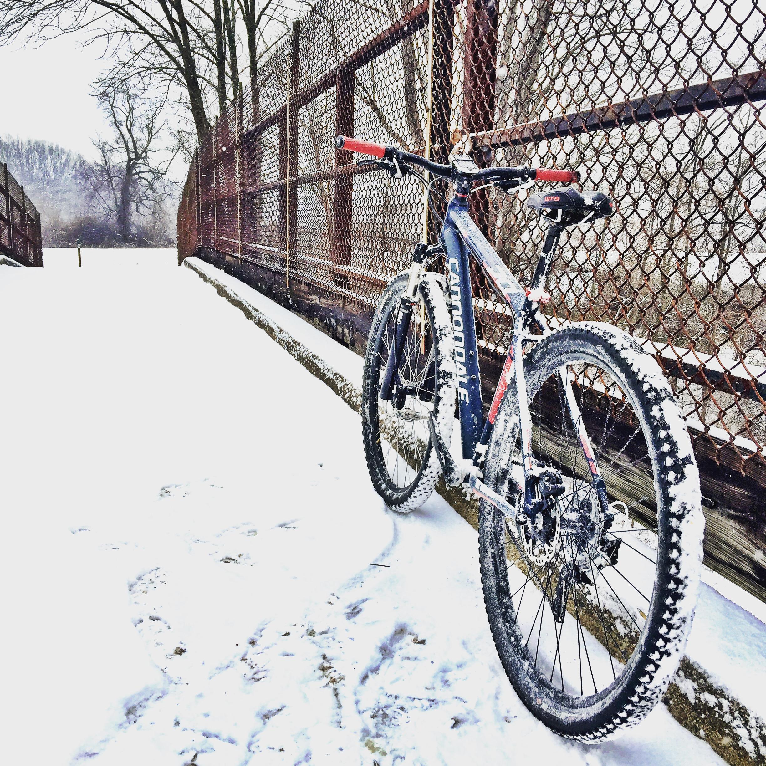 A mountain bike covered in snow rests against a weathered wooden railing on a snow-covered path. The background features a snowy landscape with bare trees and a chain-link fence. Fair Hill mountain bike trail.