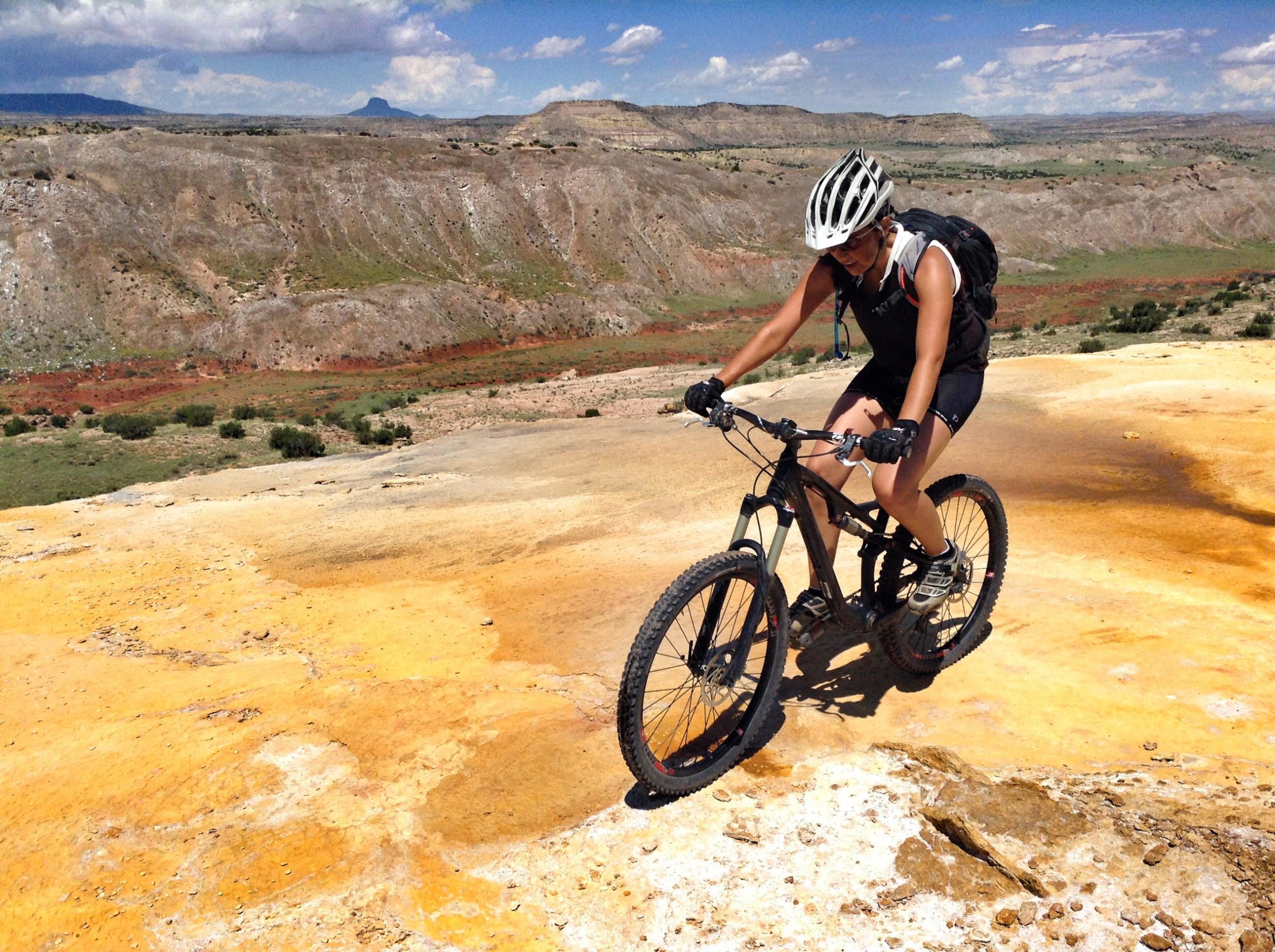 A person riding a mountain bike on a rocky, colorful terrain with a scenic backdrop of hills and clouds. The cyclist is wearing a helmet and black athletic gear, focused on navigating the challenging landscape under a sunny sky. White Ridge Bike Trails mountain bike trail.