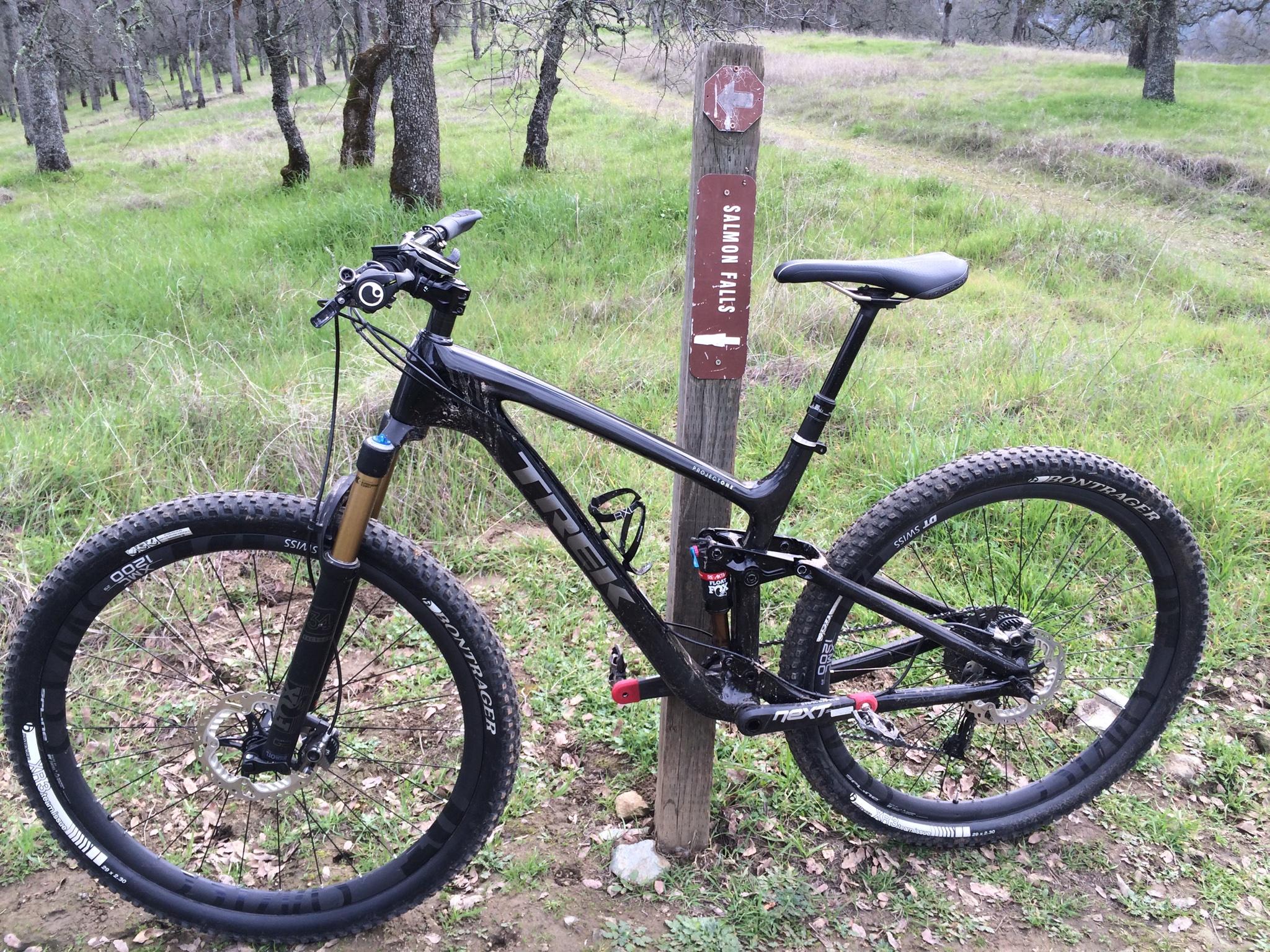 A black mountain bike parked next to a trail sign indicating "Salmon Falls," set in a grassy area surrounded by trees. Salmon Falls: Sweetwater Loop mountain bike trail.