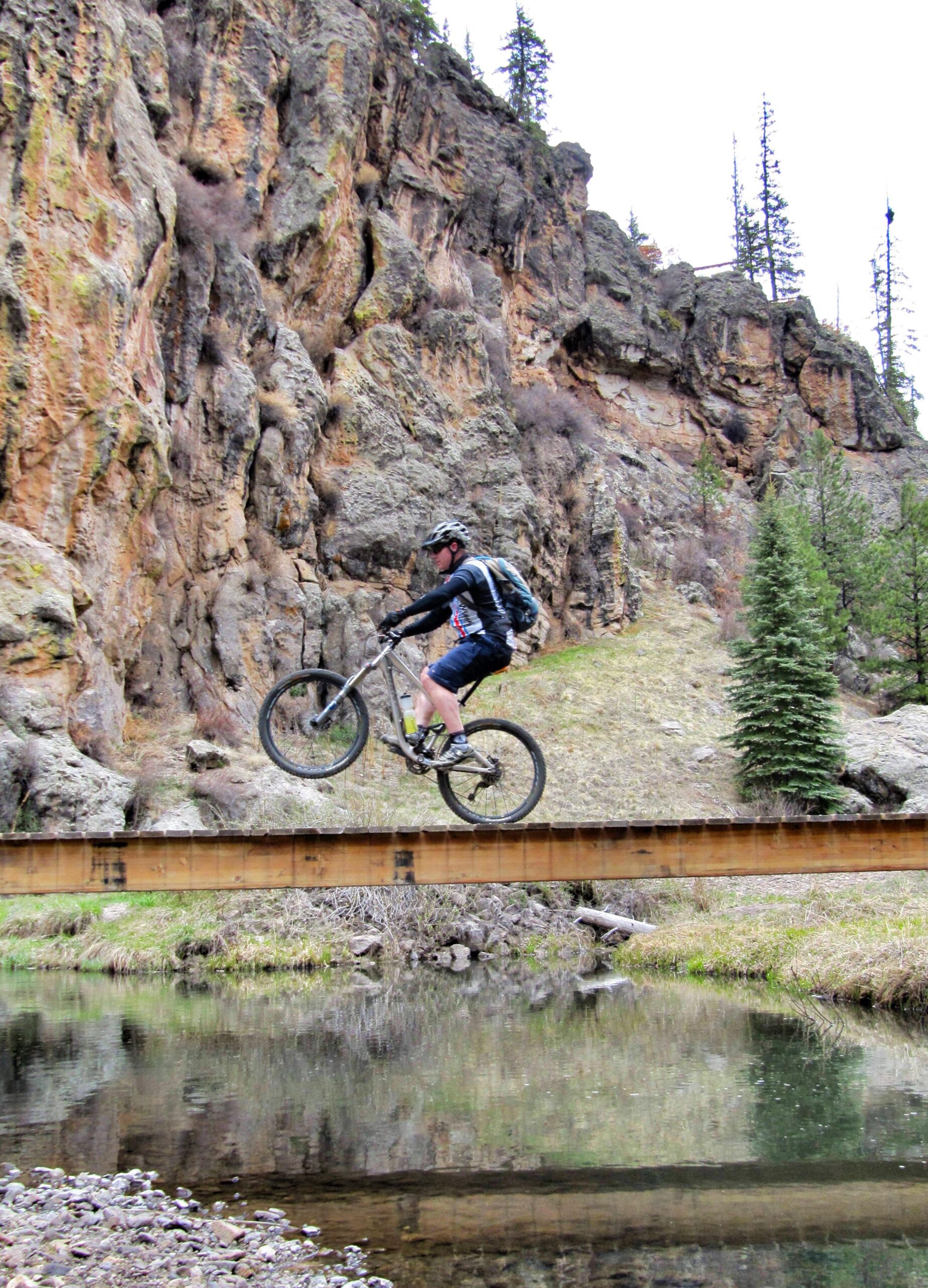 A mountain biker performs a jump over a wooden bridge, with rocky cliffs and a forested area in the background. A reflection of the biker and the bridge can be seen in the calm water below.