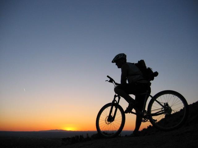 Silhouette of a cyclist on a mountain bike, positioned against a vibrant sunset sky, with soft hues of orange and blue. The rider is wearing a helmet and has a backpack, capturing a moment of outdoor adventure.