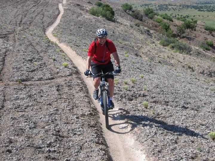 A person riding a mountain bike down a narrow dirt trail on a rocky hillside, surrounded by sparse vegetation and rolling hills in the background. The cyclist is wearing a helmet and a red shirt, with sunlight illuminating the scene. White Ridge Bike Trails mountain bike trail.