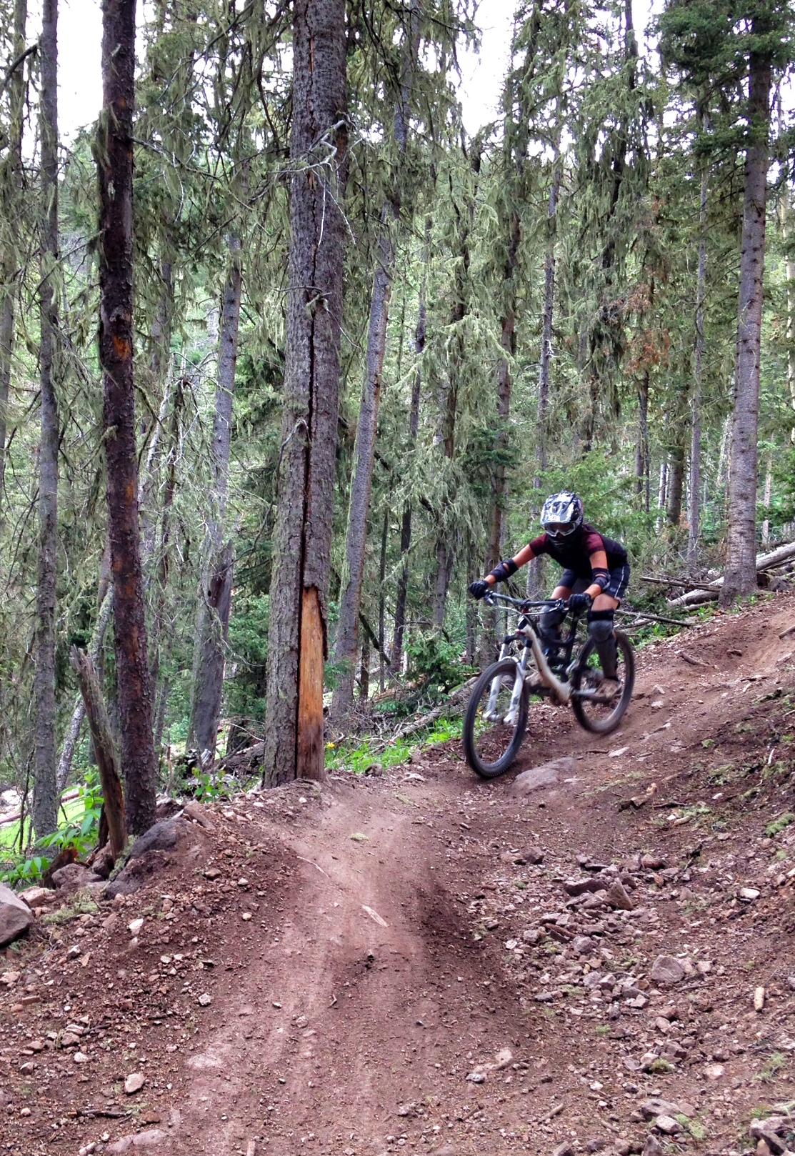 A mountain biker navigating a dirt trail through a forest, surrounded by tall trees and greenery. The cyclist is wearing a helmet and protective gear, leaning forward on the bike as they maneuver a turn on the path. Angel Fire Bike Park mountain bike trail.