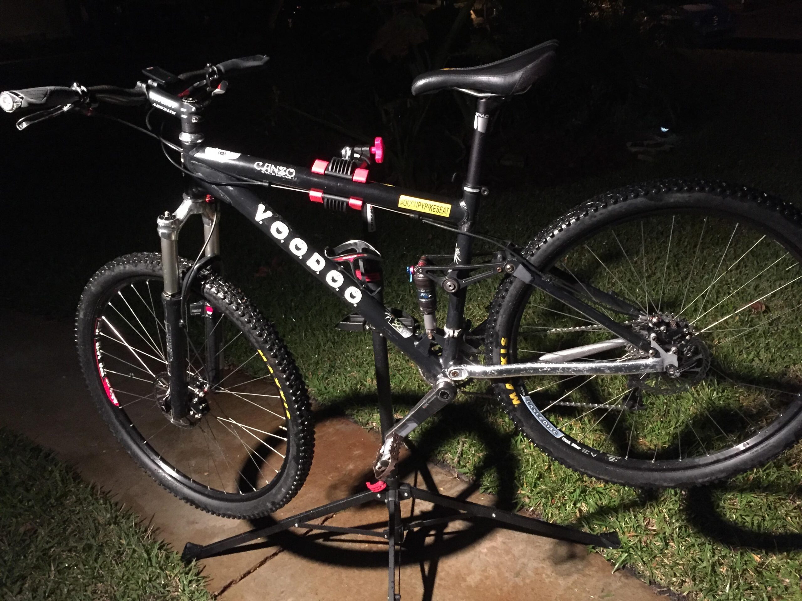 Voodoo Cycles Canzo 29er F/S: A black mountain bike is displayed on a repair stand at night, with bright lighting highlighting its features. The bike has thick, knobby tires and a sturdy frame with branding that includes "Voodoo" and "Canzos." The handlebars are equipped with grips and controls, and the bike appears well-maintained with a shiny metal drivetrain. The background includes grass and hints of an outdoor environment, suggesting a casual setting.