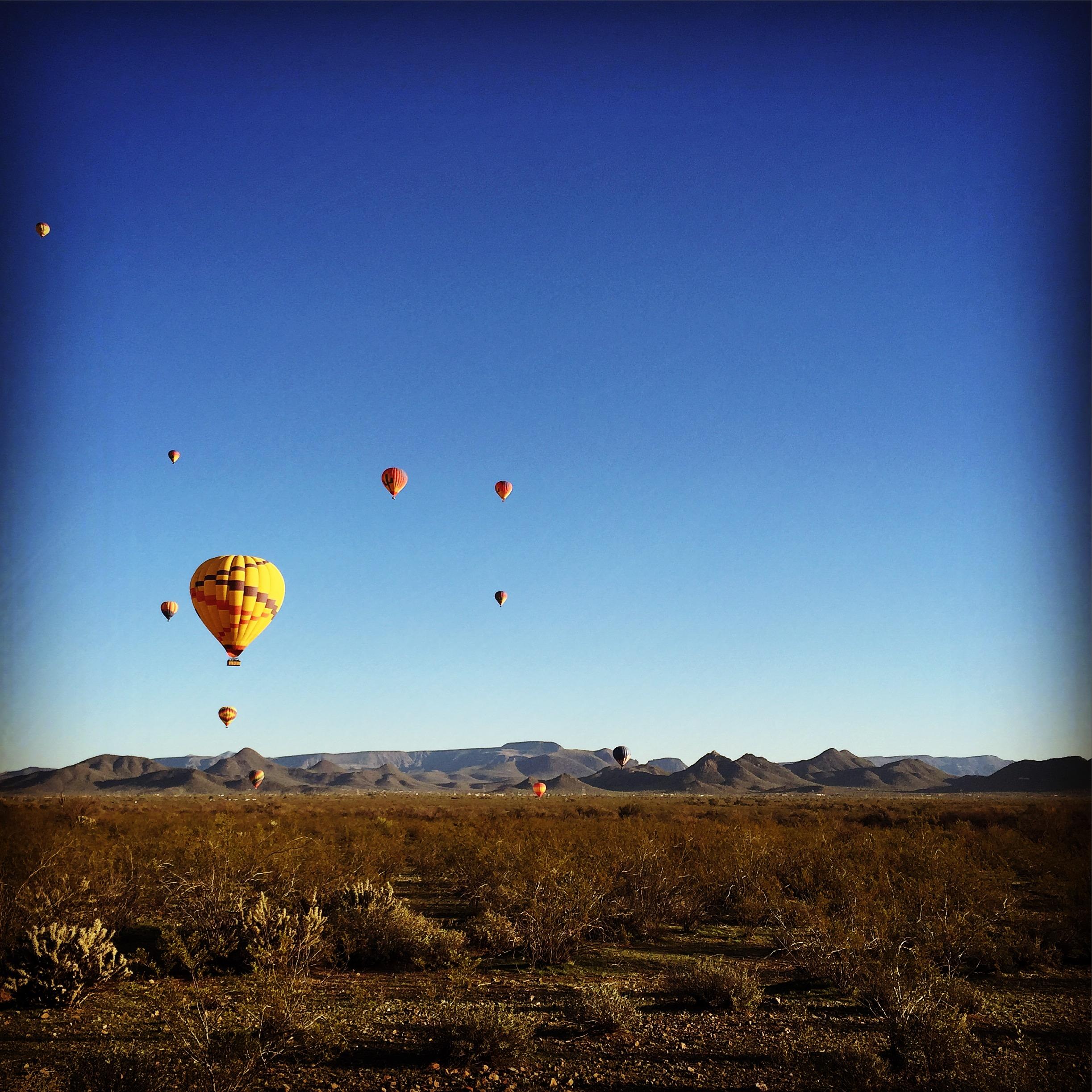 A scenic view of colorful hot air balloons ascending in a clear blue sky over a vast desert landscape with distant mountains and sparse vegetation. Sonoran Trail mountain bike trail.