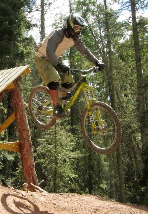 A mountain biker mid-air while jumping off a wooden ramp in a forested area. The cyclist is wearing a helmet and protective gear, with a focus on showcasing the action and excitement of mountain biking. Angel Fire Bike Park mountain bike trail.