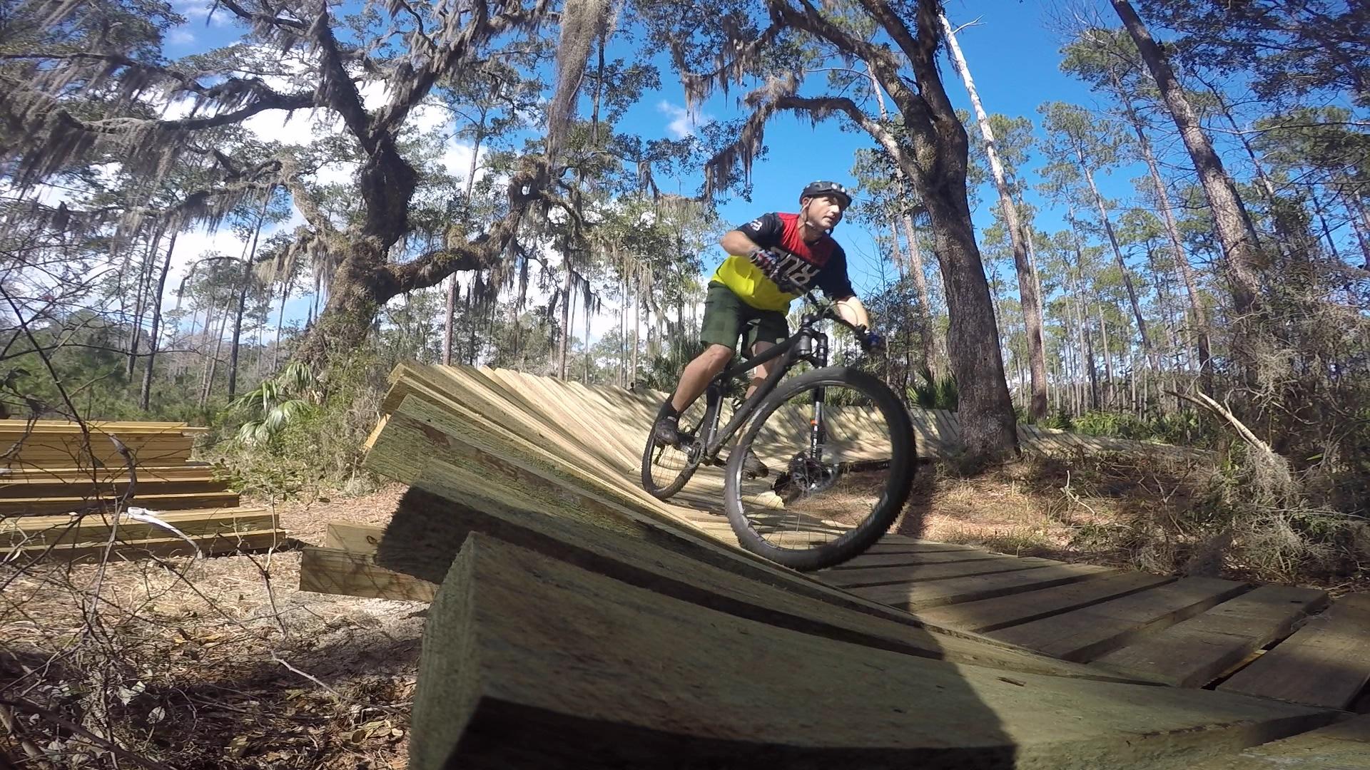 A person riding a mountain bike on a wooden ramp in a forested area, surrounded by tall trees and moss. The cyclist is wearing a helmet and colorful cycling gear, navigating the smooth, curved wooden surface under a bright blue sky. Nocatee mountain bike trail.