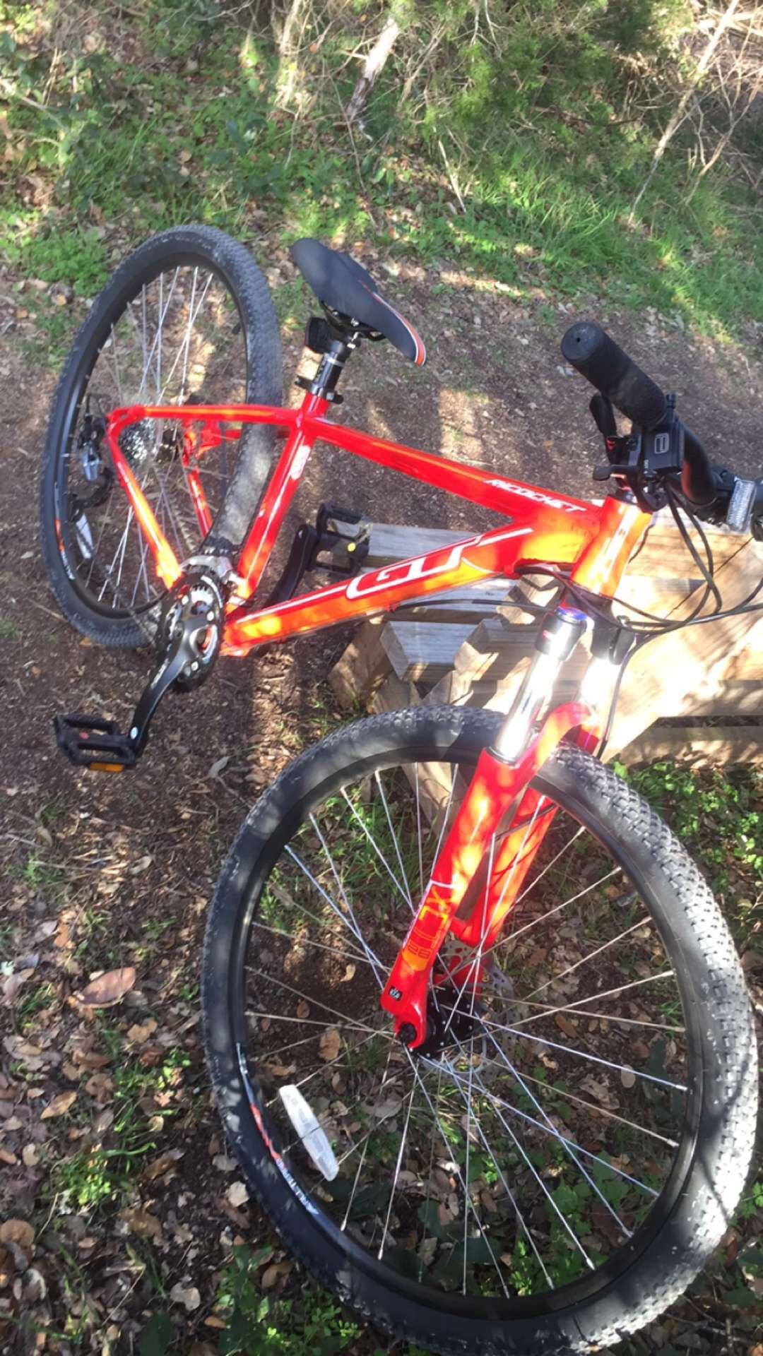 GT Ricochet: A bright red mountain bike lying on a dirt path surrounded by grass and small leaves. The bike features thick tires and a modern design, with visible gears and pedals. In the background, there are bushes and trees, indicating a natural outdoor setting.