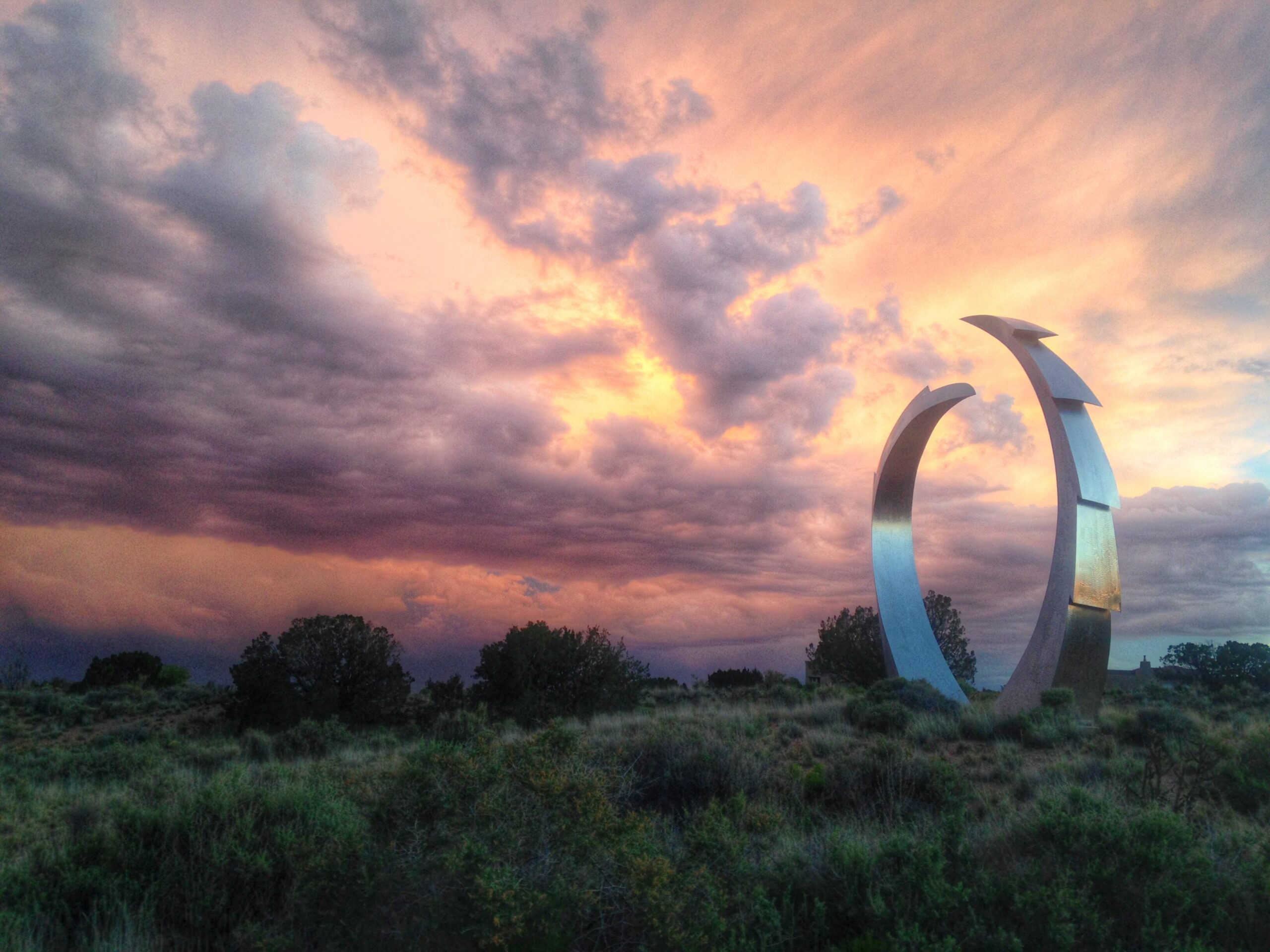 A modern metallic sculpture resembling two intersecting rings, set against a dramatic sky filled with clouds in shades of purple and orange during sunset. Lush green vegetation and trees are visible in the foreground. Parkway Fatbike trail mountain bike trail.