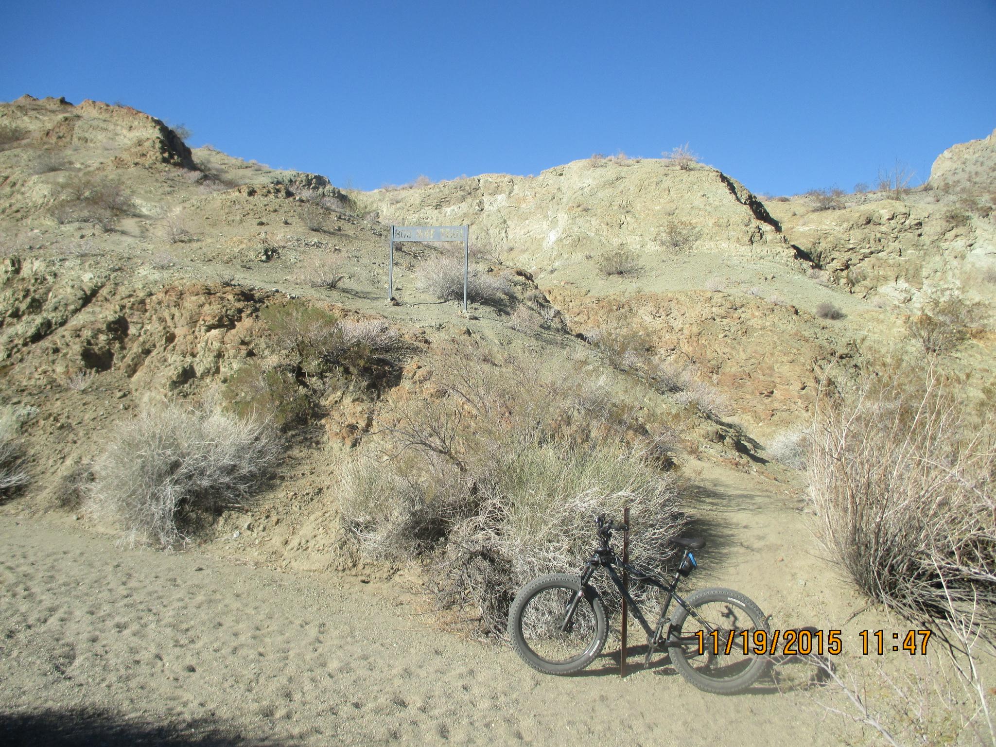 Rocky Mountain Blizzard: A black mountain bike is resting on a sandy trail in a rocky landscape, with sparse vegetation and brush surrounding it. The path leads up a hill, and a metal signpost can be seen in the background, partially obscured by the terrain. The sky above is clear and blue.