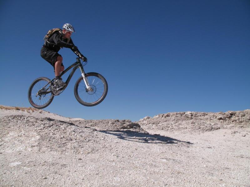 A mountain biker in mid-air, performing a jump over a rocky terrain against a clear blue sky. The rider is wearing a helmet and sports gear, with a backpack visible. White Ridge Bike Trails mountain bike trail.