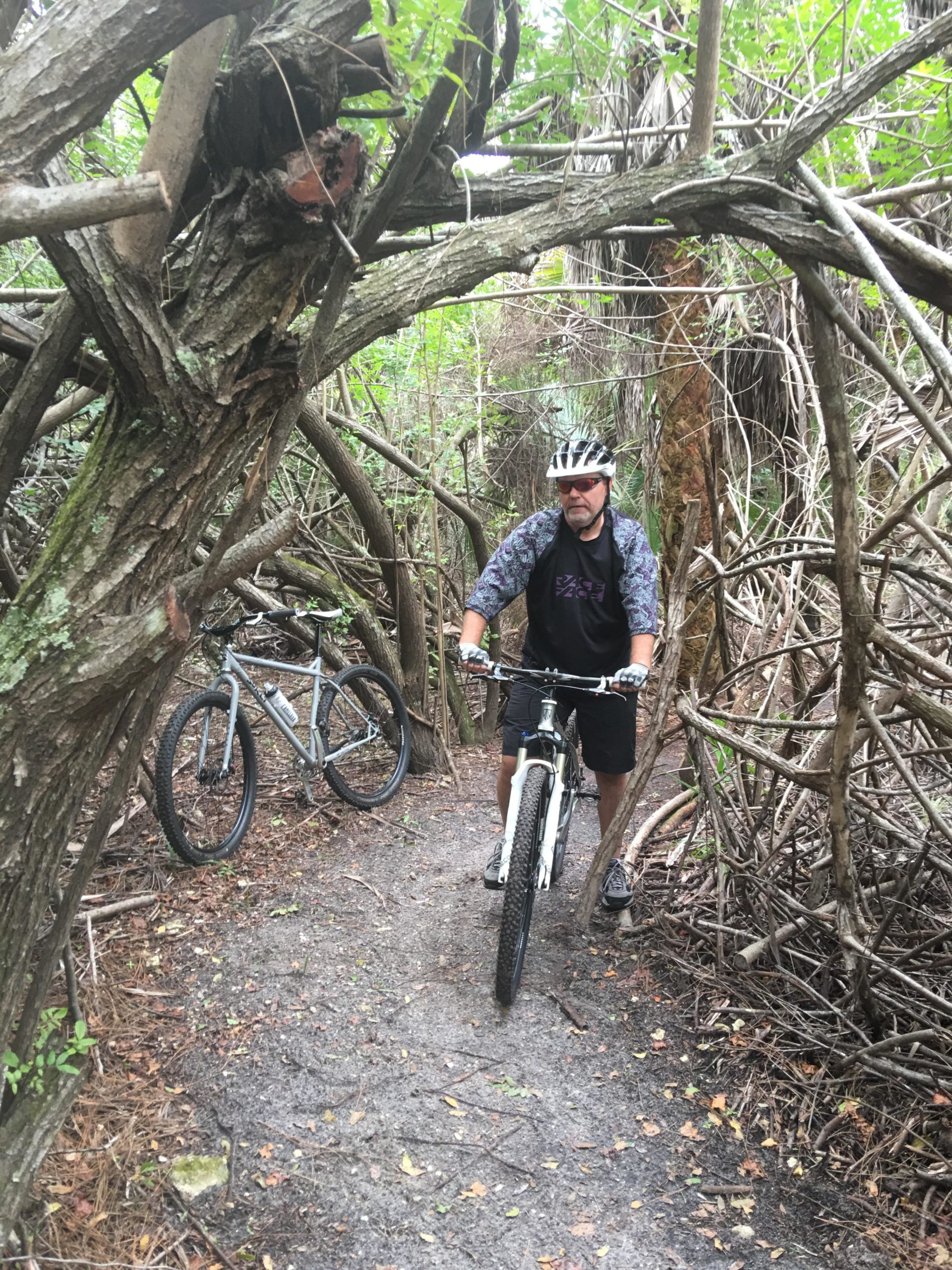 A mountain biker navigates a narrow, overgrown trail surrounded by thick vegetation and branches, with a second bicycle parked nearby. The cyclist wears a helmet and casual biking attire, showcasing a challenging and rugged outdoor environment. Hillsboro Express mountain bike trail.