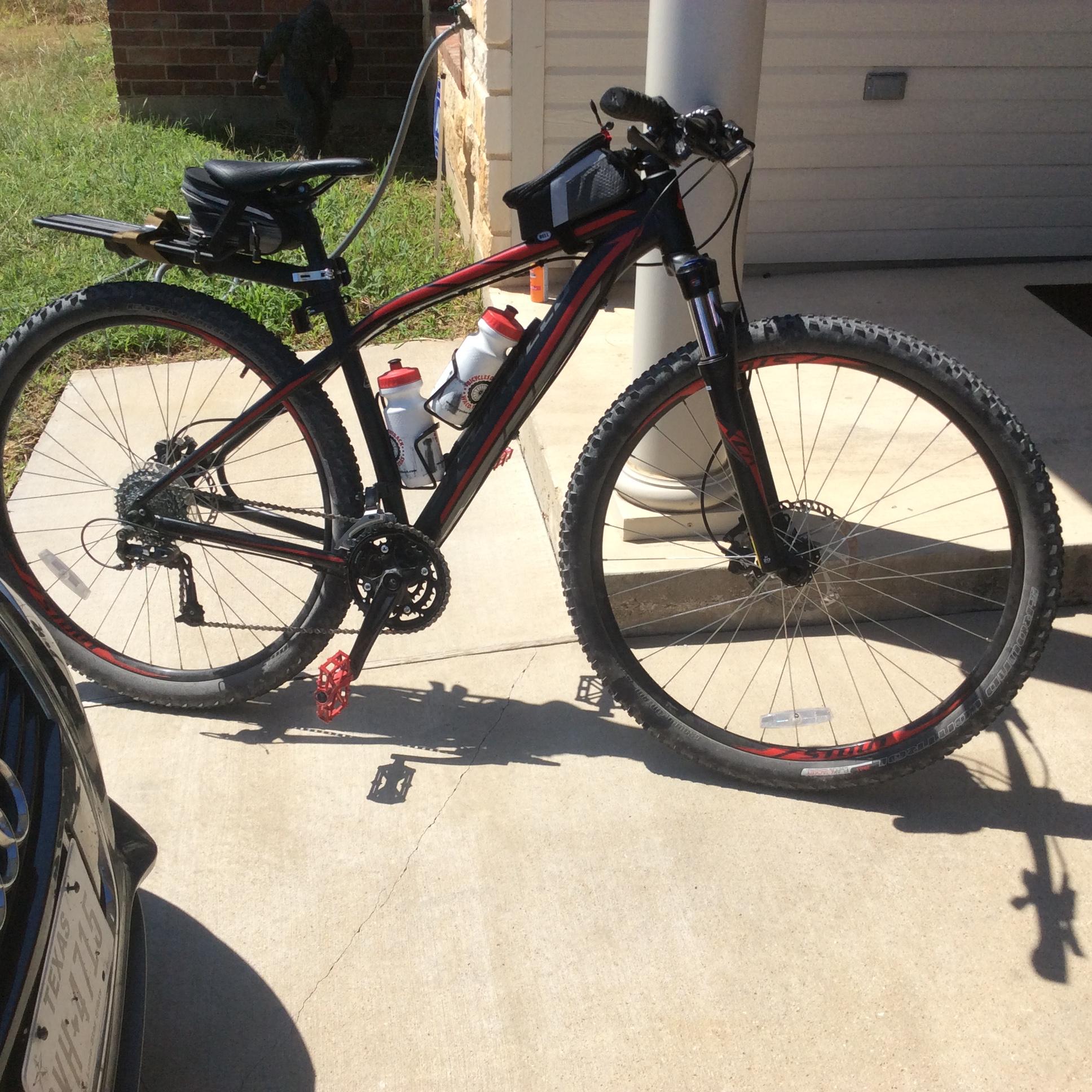 Specialized Rockhopper 29: A black and red mountain bike is parked on a concrete surface, with two water bottles mounted on the frame. The bike has large, knobby tires suitable for off-road riding, and a small black bag attached to the back. In the background, there is a patch of grass and part of a house.