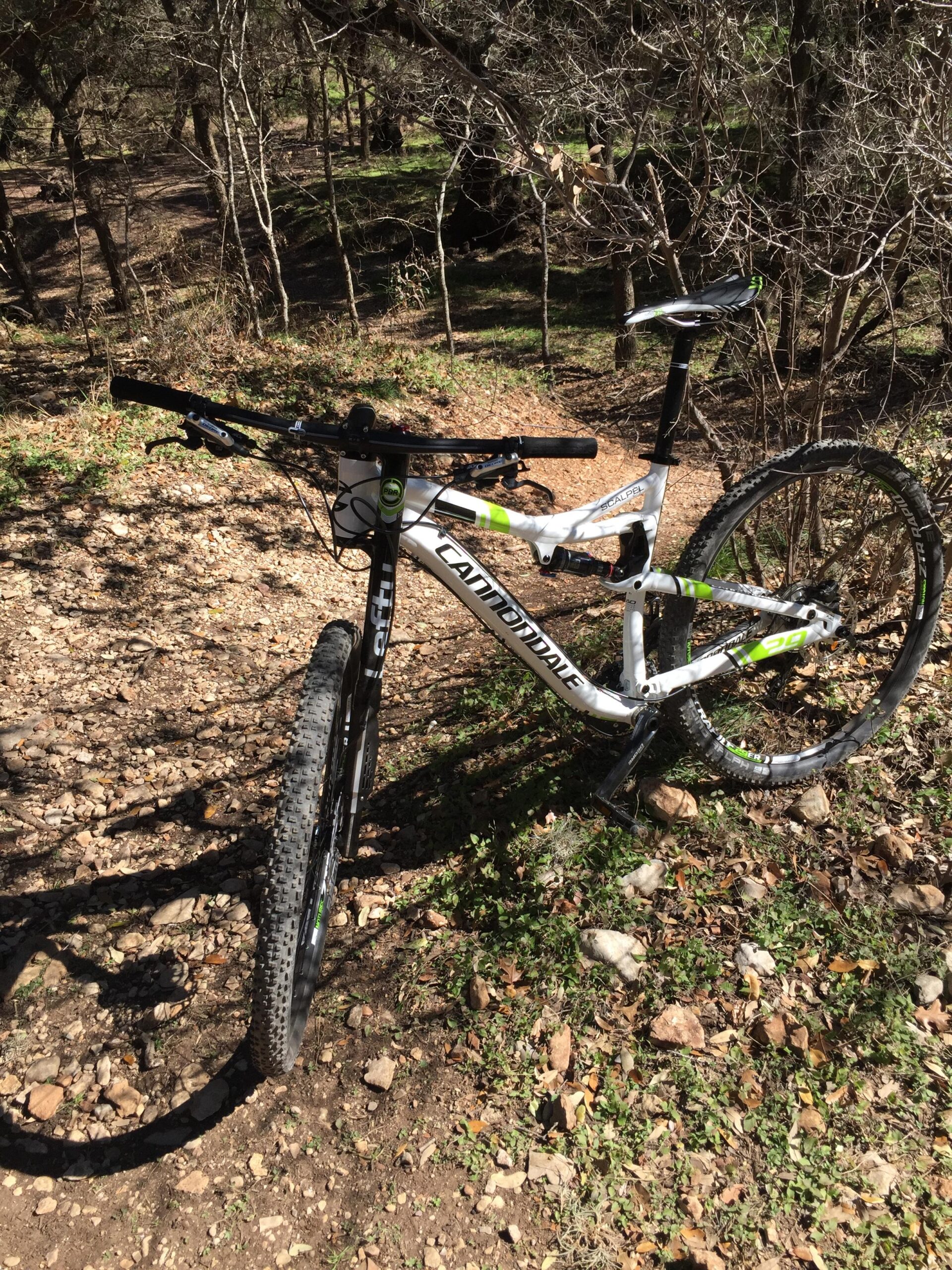 A mountain bike positioned on a rocky trail surrounded by trees, with a clear view of its frame and tires. The bike features a white and green color scheme and is resting on a natural outdoor surface with dirt and scattered stones.