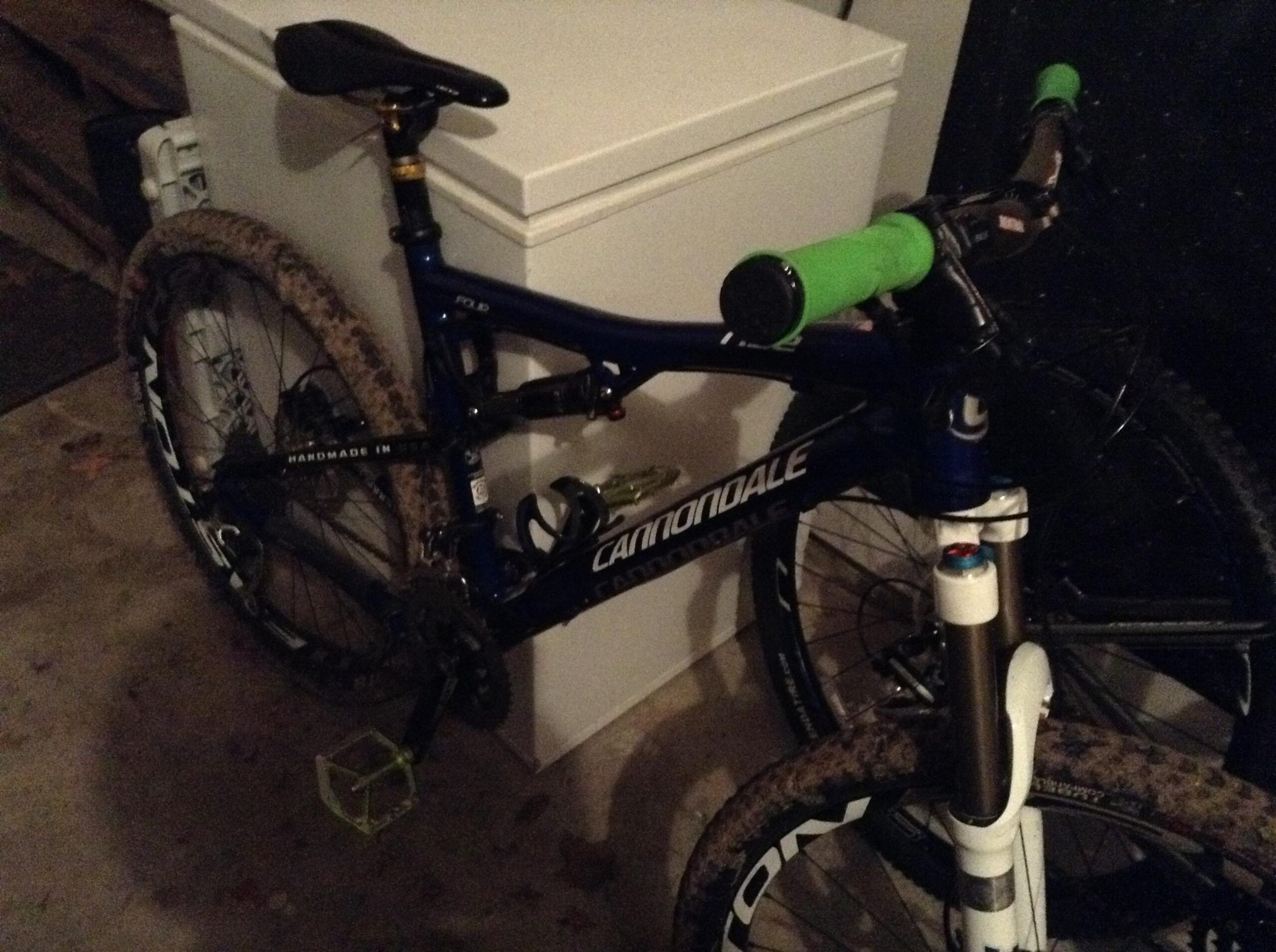 Cannondale Rize 4: A blue Cannondale mountain bike with green handlebar grips, leaning against a white chest freezer. The bike features muddy tires, a black crankset with white pedals, and visible gears and suspension components. The background shows a dark garage setting.
