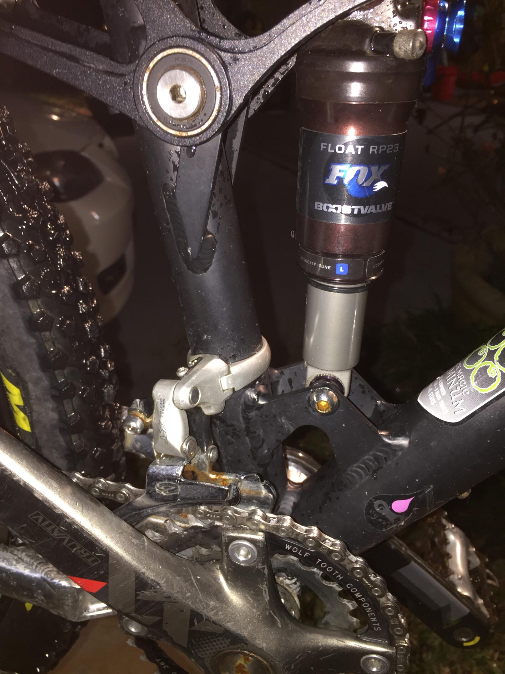 Voodoo Cycles Canzo 29er F/S: Close-up view of a mountain bike's rear suspension, featuring a FOX Float RP23 shock absorber, muddy and wet components, and a visible chain and derailleur. The setting appears to be outdoors at night.