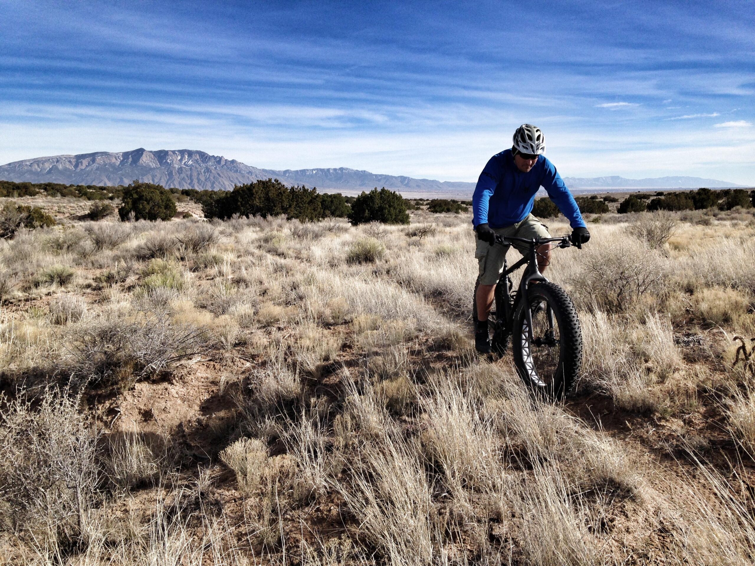 A person riding a fat tire bike on a rugged, grassy trail with mountains in the background. The cyclist is wearing a blue long-sleeve shirt and a helmet, focused on navigating the terrain. The landscape features sparse vegetation and a clear blue sky. Parkway Fatbike trail mountain bike trail.
