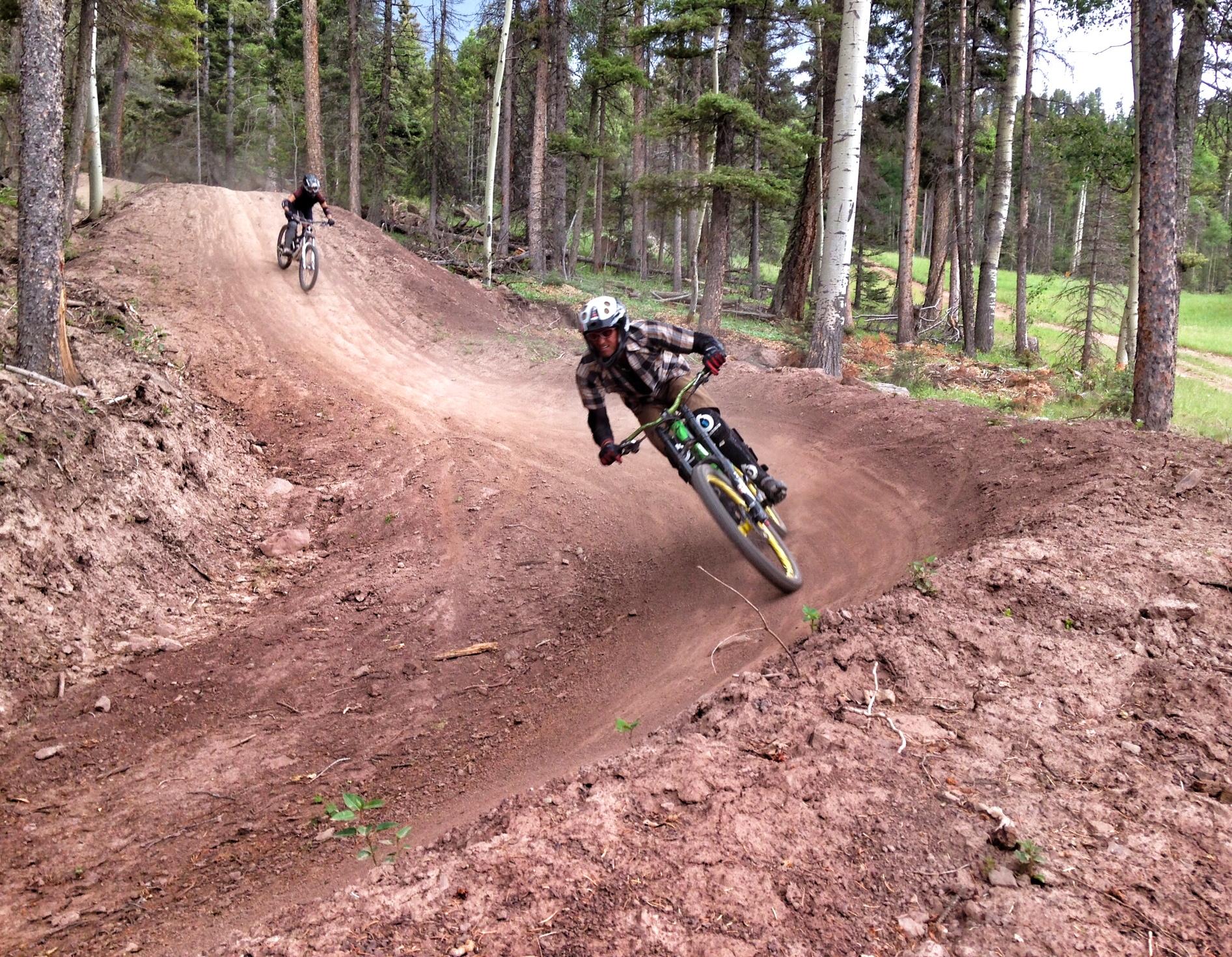 Two mountain bikers navigating a dirt trail in a forested area, one taking a sharp turn while the other rides in the background. The scene captures the excitement of outdoor biking amidst trees and a clear sky. Angel Fire Bike Park mountain bike trail.