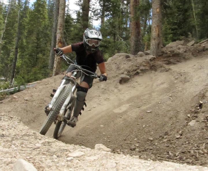 A person wearing a helmet and protective gear is riding a mountain bike on a dirt trail surrounded by trees. The rider is leaning into a turn, kicking up dust and gravel as they navigate the path. The scene captures a sense of adventure and outdoor activity. Angel Fire Bike Park mountain bike trail.