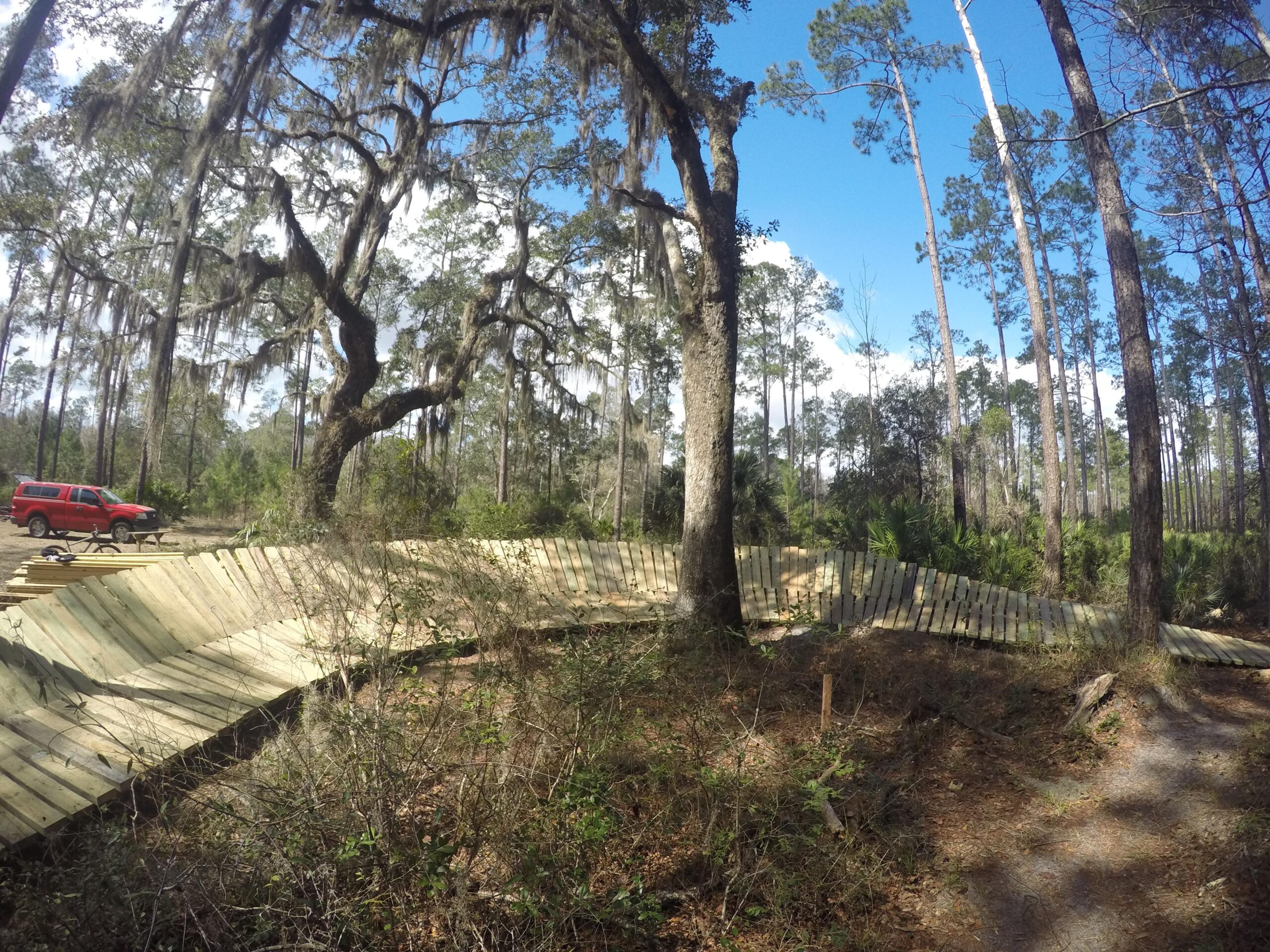 A winding wooden path surrounded by tall trees and lush vegetation, with a red vehicle parked nearby. The scene showcases a sunny day in a forested area, highlighting the natural beauty of the landscape. Nocatee mountain bike trail.