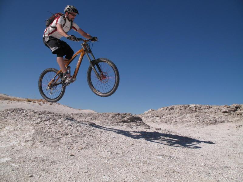 A mountain biker in a helmet and cycling gear is performing a jump over a rocky terrain, with clear blue skies in the background. The bike's front wheel is elevated off the ground as the rider maintains a focused posture in mid-air. Shadows cast on the ground enhance the sense of motion and excitement. White Ridge Bike Trails mountain bike trail.