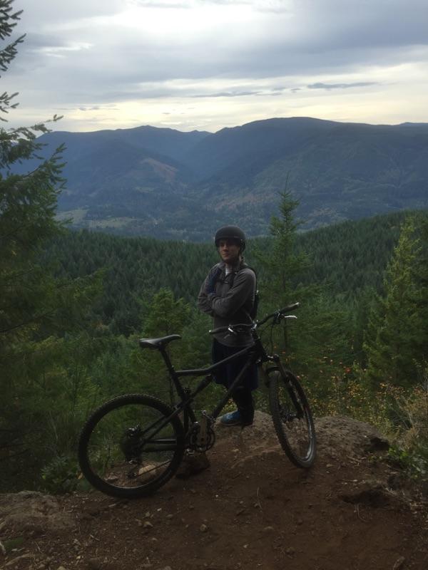 Santa Cruz Blur: A person wearing a helmet and a gray jacket stands with arms crossed next to a mountain bike on a rocky outcrop, overlooking a lush green valley and distant mountains under a cloudy sky.