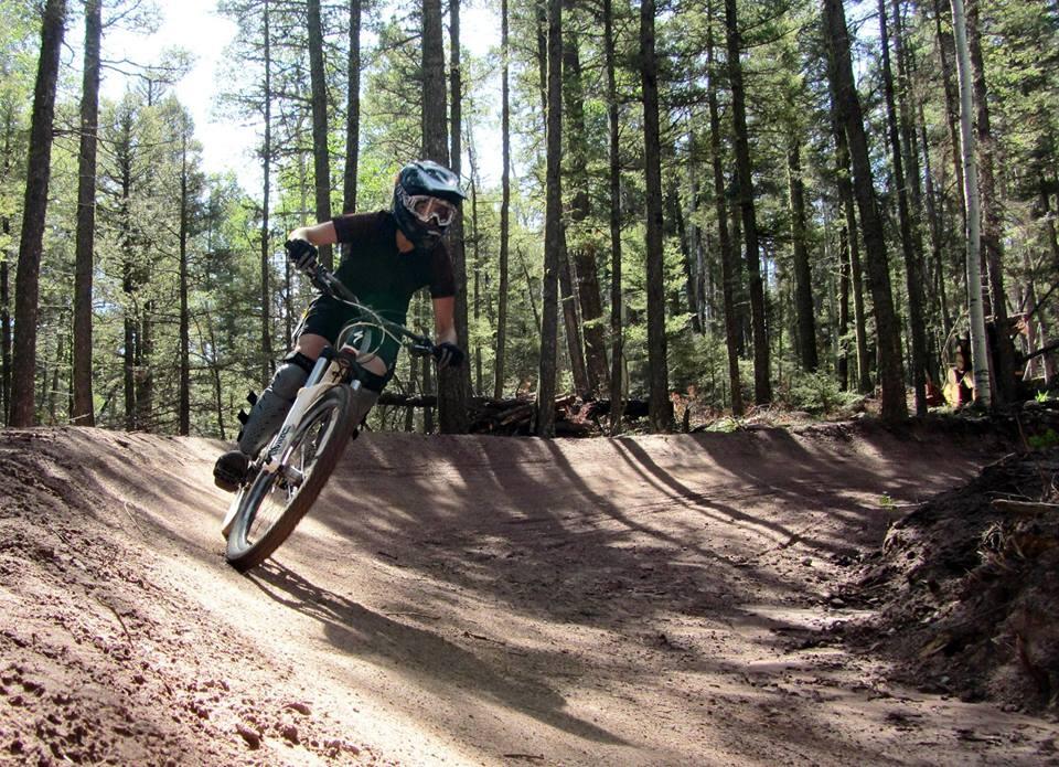 A mountain biker wearing protective gear leans into a turn on a dirt trail surrounded by tall trees in a forest. The sunlight filters through the leaves, creating shadows on the trail. Angel Fire Bike Park mountain bike trail.
