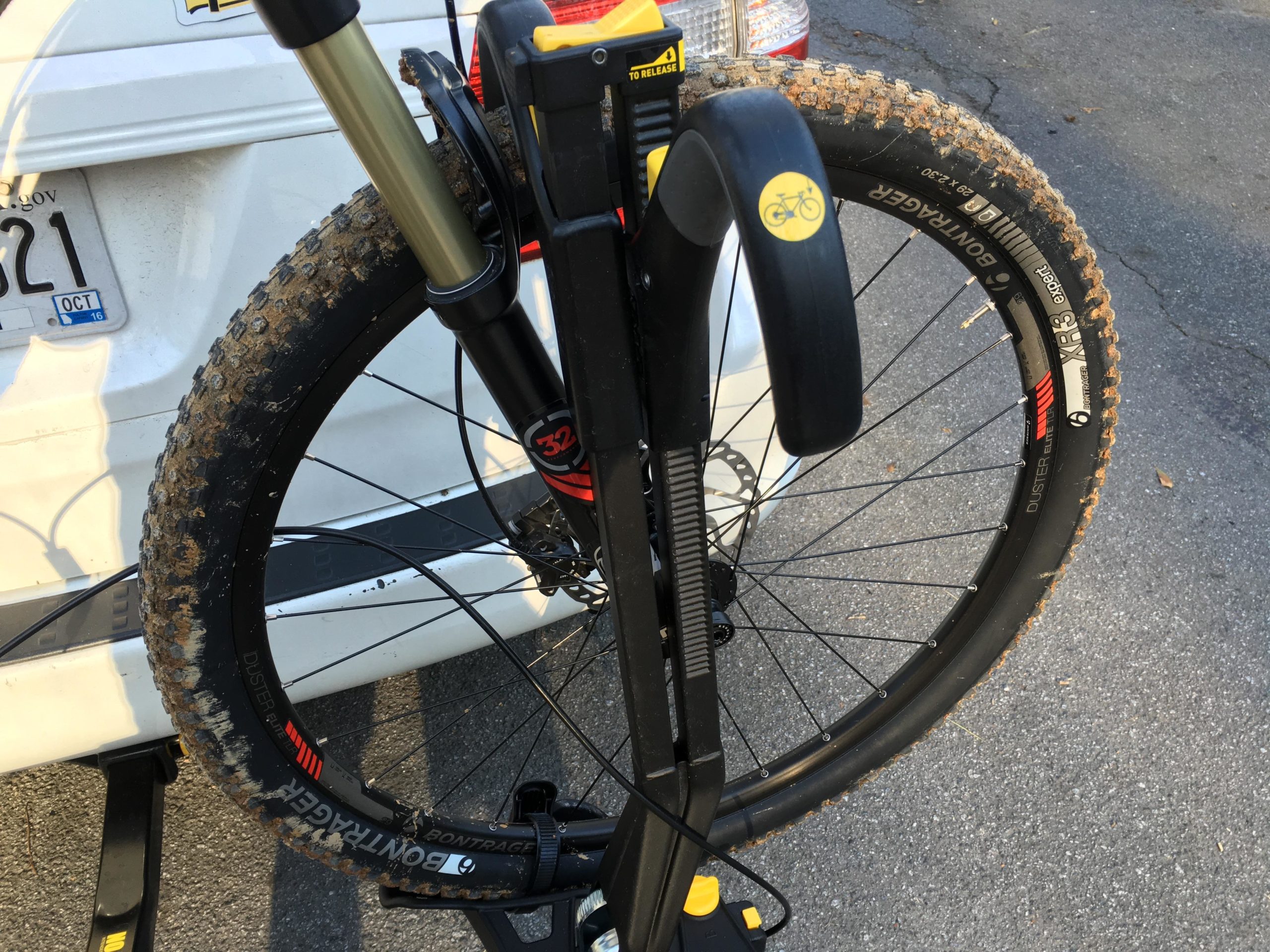 Close-up view of a mountain bike's front fork and tire mounted on a bike rack. The tire shows signs of dirt and use, indicating recent off-road activity. In the background, part of a vehicle is visible with a license plate. Sope Creek mountain bike trail.