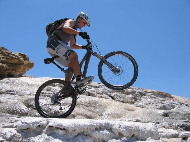 A mountain biker performing a stunt on rocky terrain, lifting the front wheel off the ground while wearing a helmet and athletic gear, under a clear blue sky. White Ridge Bike Trails mountain bike trail.