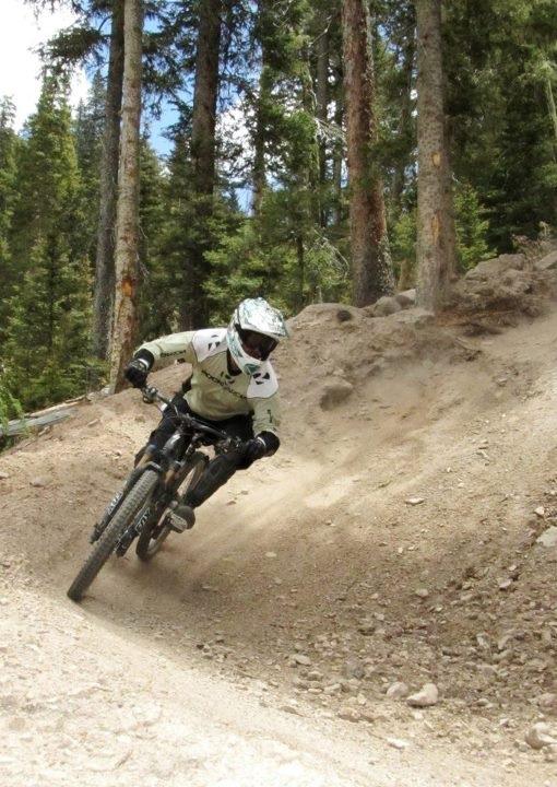 A mountain biker leaned into a turn on a dirt trail surrounded by trees, with dust rising from the ground. The rider is wearing a full-face helmet and protective gear, and appears to be in mid-motion, navigating the curve on a mountain bike. Angel Fire Bike Park mountain bike trail.