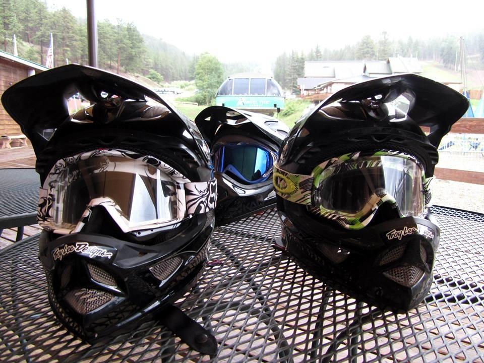 Three black motocross helmets with different designs sit on a metal table outdoors. Each helmet features distinct colored goggles, with one adorned in a green and black pattern. In the background, there are blurred images of trees and buildings in a mountainous setting. The atmosphere appears misty, suggesting a cool, overcast day. Angel Fire Bike Park mountain bike trail.