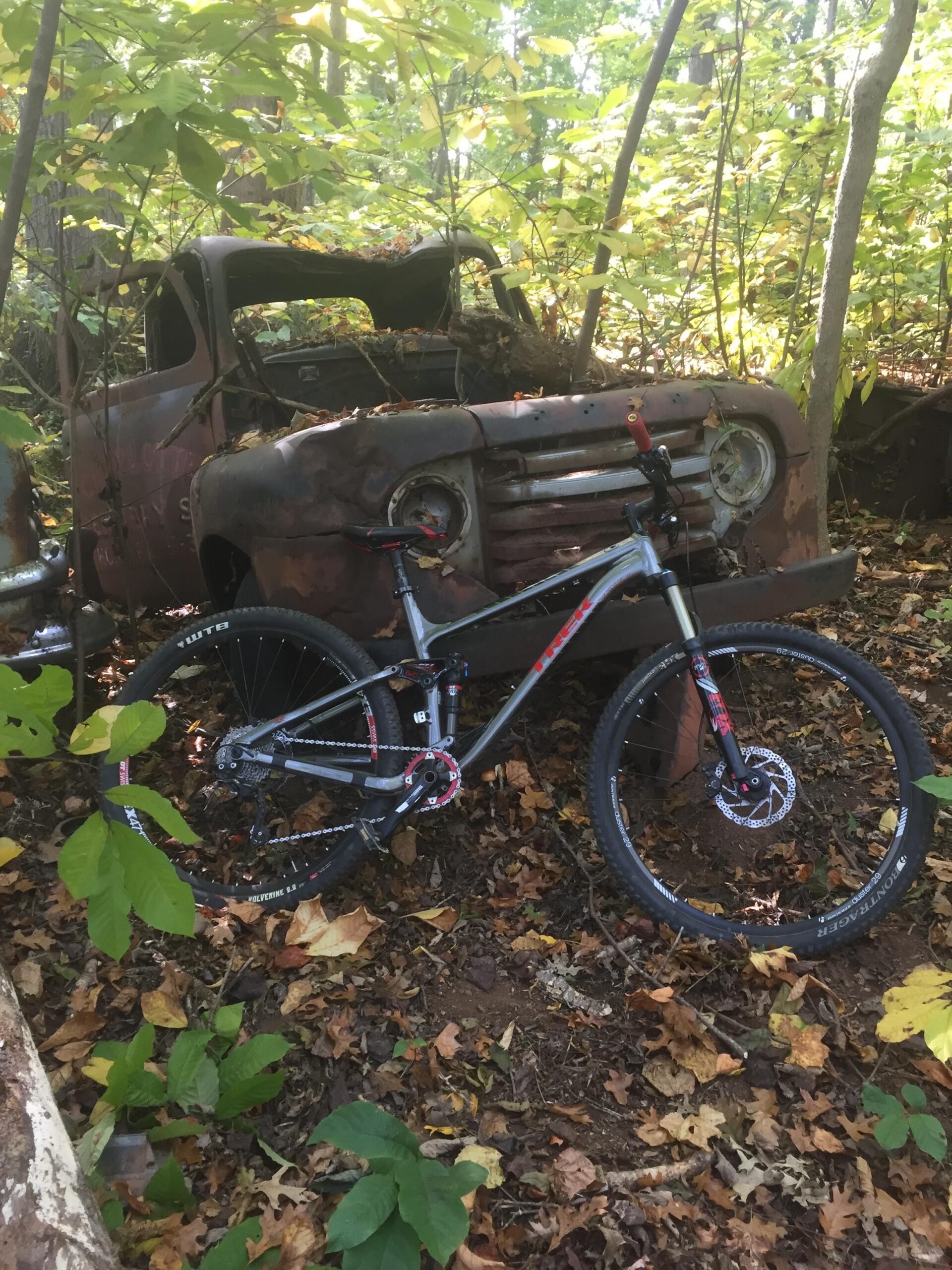 Trek Fuel EX 8 29: A mountain bike rests against an old, rusted truck partially hidden in a forested area. The ground is covered with fallen leaves, and greenery surrounds the scene, creating a blend of nature and vintage remnants.