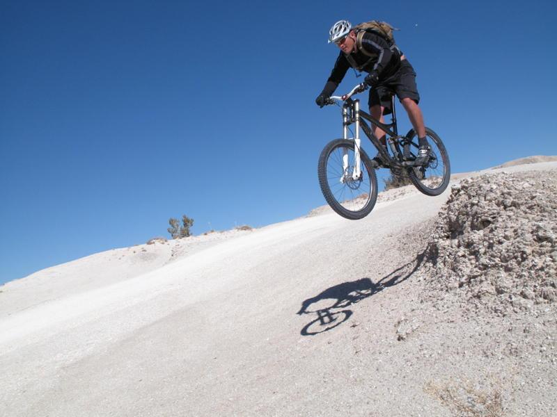 A mountain biker in mid-air performing a jump over a dirt mound on a sunny day, with a clear blue sky in the background. White Ridge Bike Trails mountain bike trail.