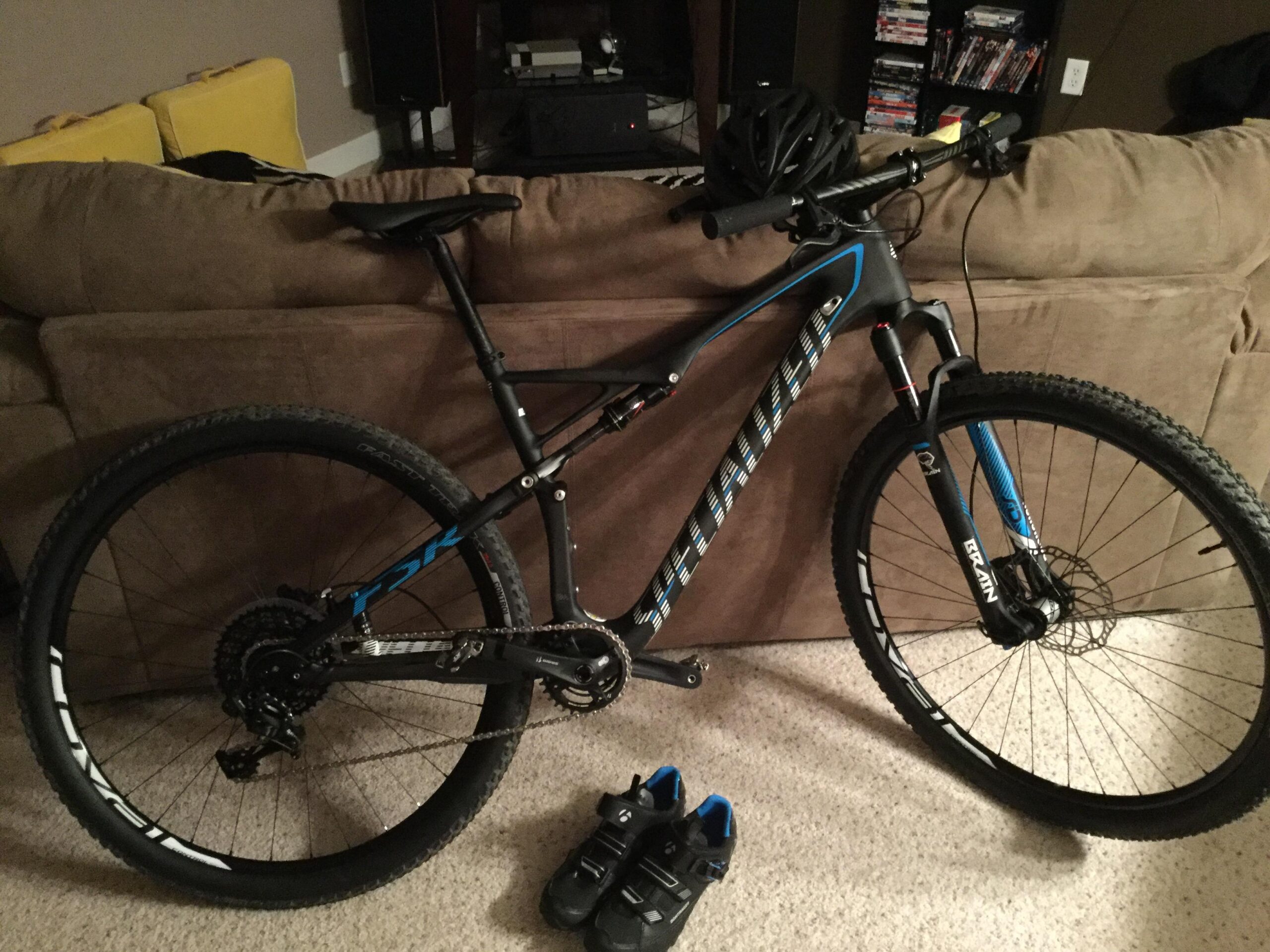 Specialized Epic Expert Carbon World Cup: A mountain bike leaning against a couch in a living room, accompanied by a helmet on the seat and a pair of cycling shoes on the floor. The bike features a black and blue frame, with prominent tires designed for off-road riding.