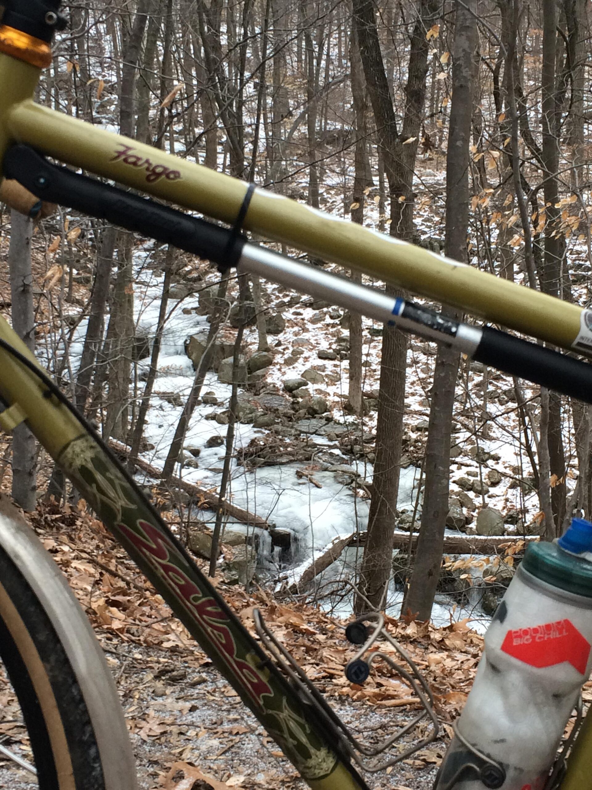 A close-up view of a bike frame in a wooded area, with fallen leaves scattered on the ground. In the background, a partially frozen stream flows among rocks and trees, showcasing a serene winter landscape. The bike has a water bottle mounted on it, emphasizing an outdoor adventure setting. Belmont Plateau mountain bike trail.