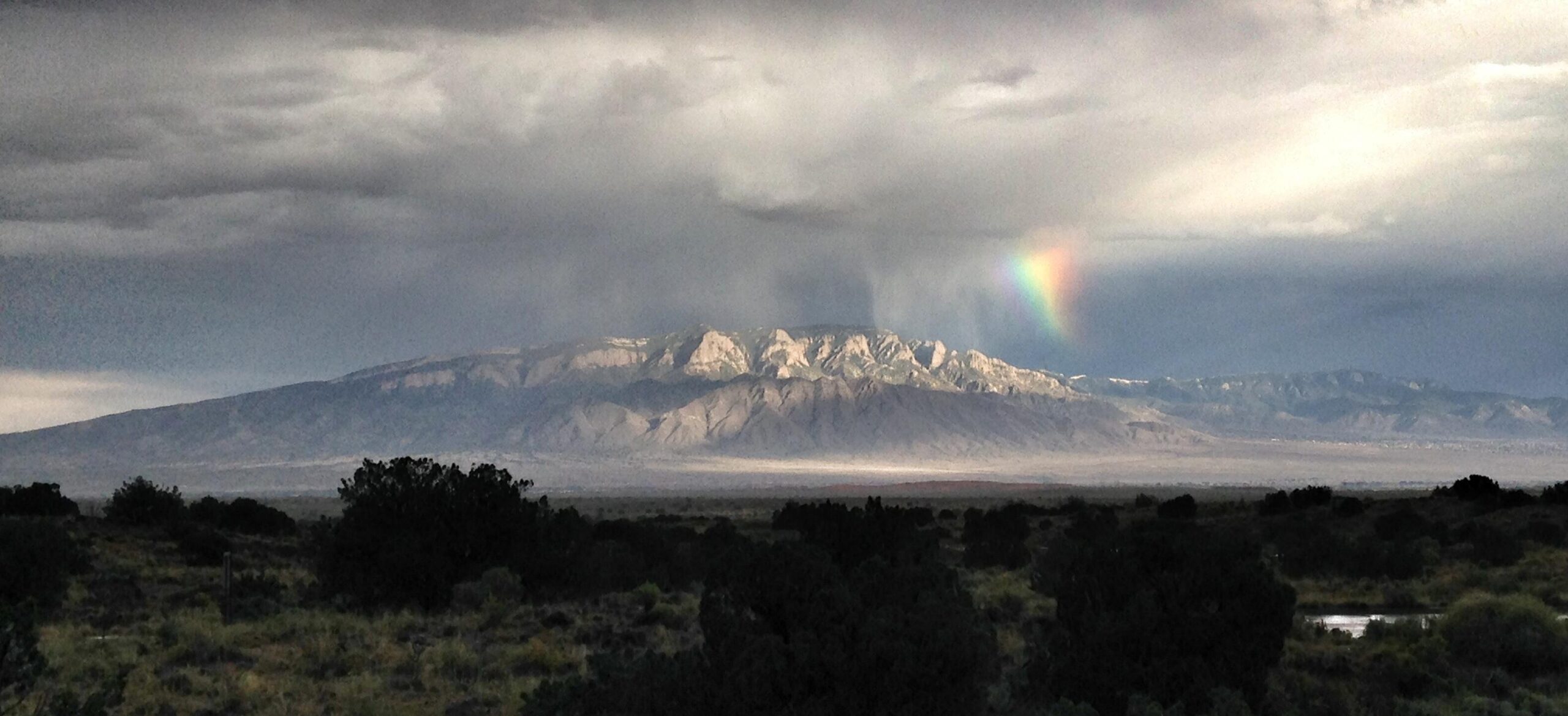 A distant mountain range under a cloudy sky, with a light rain shower visible. A faint rainbow appears to the right of the mountain, adding a splash of color to the otherwise muted scene. The foreground features sparse vegetation typical of a desert landscape. Parkway Fatbike trail mountain bike trail.