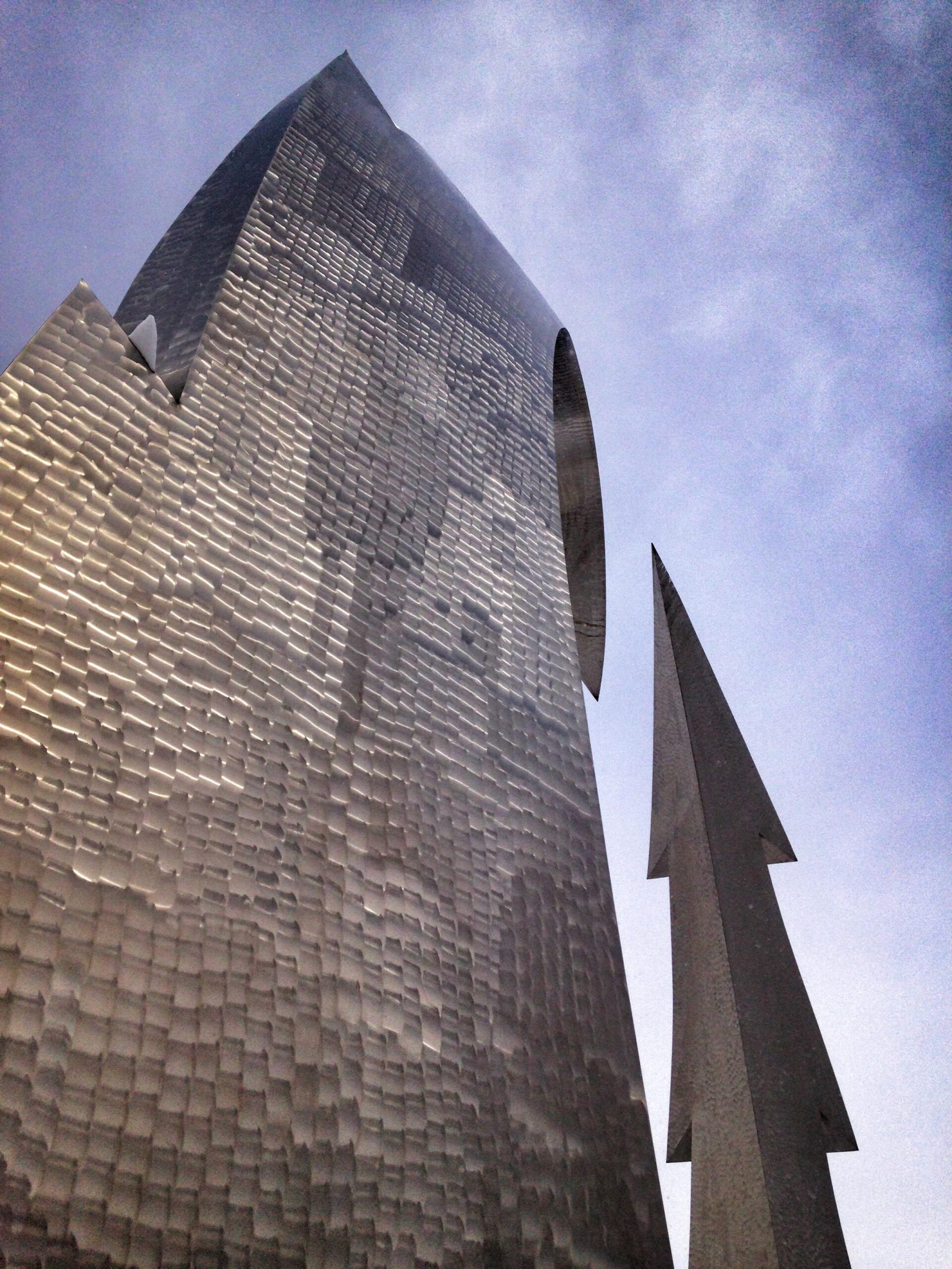 A tall, abstract metallic sculpture featuring textured surfaces and reflective elements, set against a cloudy sky. The sculpture includes a pointed structure resembling a tree, adding to its artistic presence. Parkway Fatbike trail mountain bike trail.