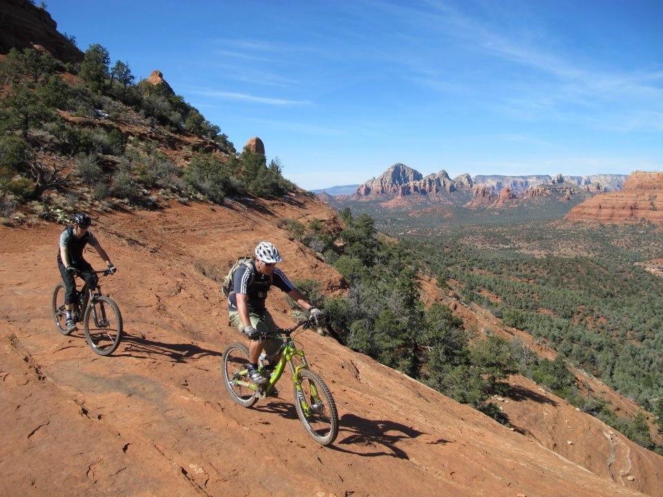 Two mountain bikers navigate a rocky trail in a desert landscape, surrounded by lush green trees and distant red rock formations under a clear blue sky. Bell Rock Area Trails mountain bike trail.