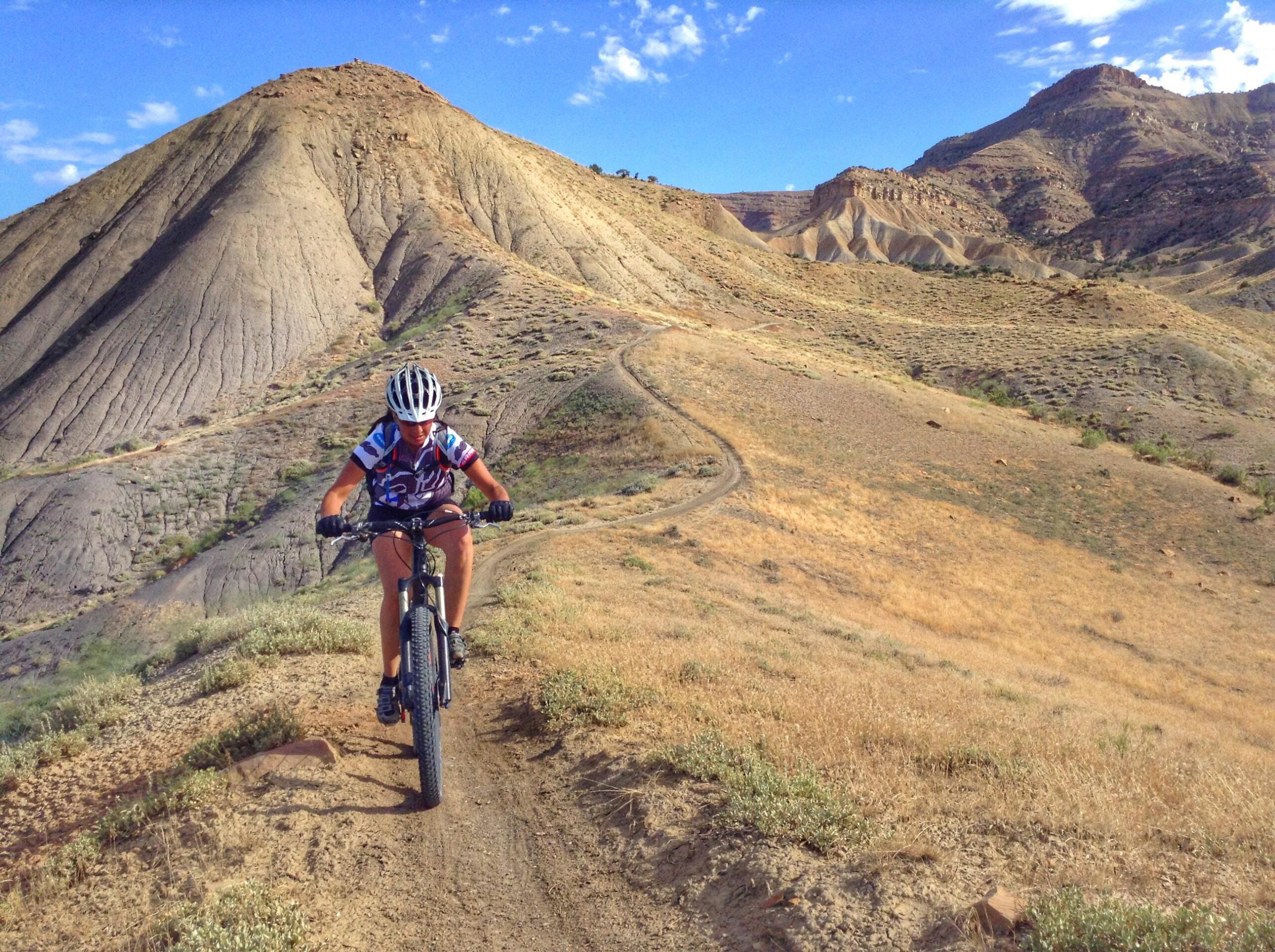 A person riding a mountain bike along a narrow trail on a rocky hillside, with arid, uneven terrain and mountains in the background under a blue sky. 18 Road Trails / North Fruita Desert mountain bike trail.