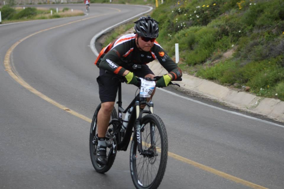 Giant Trance 27.5 2: A cyclist wearing a black and orange jersey, black shorts, and a helmet rides a mountain bike on a winding road. The cyclist is focused and in motion, with green hills and wildflowers in the background. A race number is visible on the cyclist's jersey.