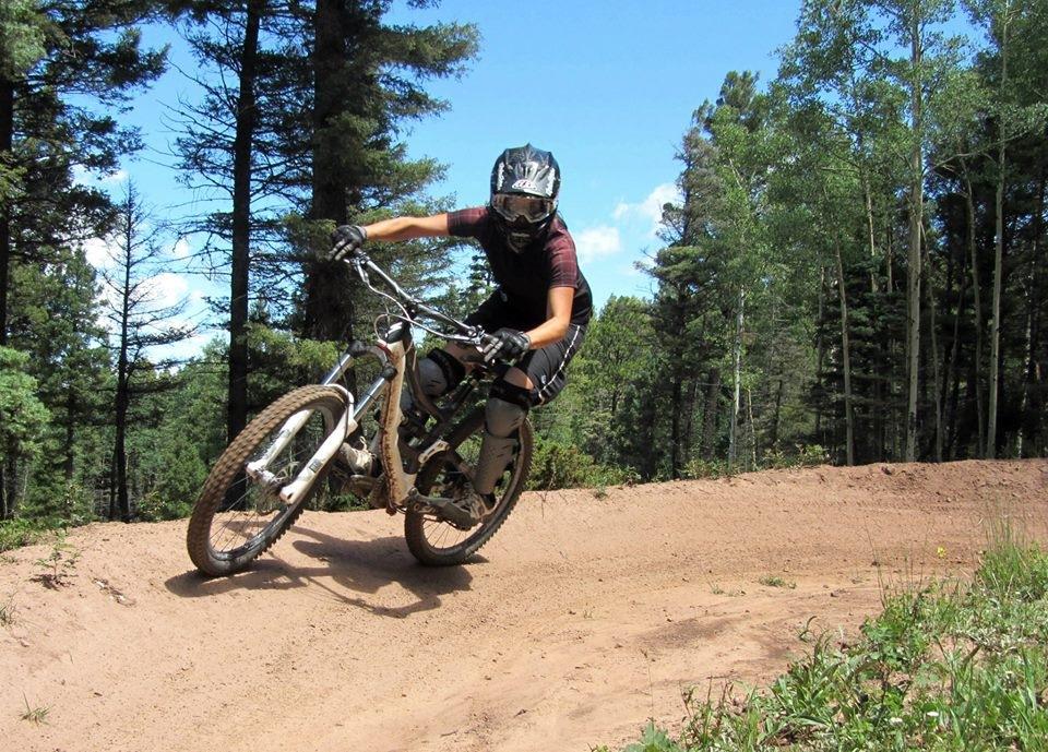 A mountain biker in protective gear navigates a dirt trail, leaning into a turn surrounded by trees under a clear blue sky. Angel Fire Bike Park mountain bike trail.