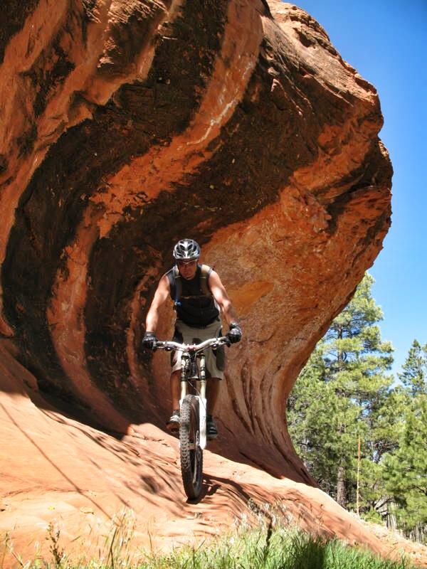A mountain biker maneuvering down a steep, rocky slope beneath a large, curved rock formation, surrounded by greenery and blue sky. High Desert Trail System mountain bike trail.
