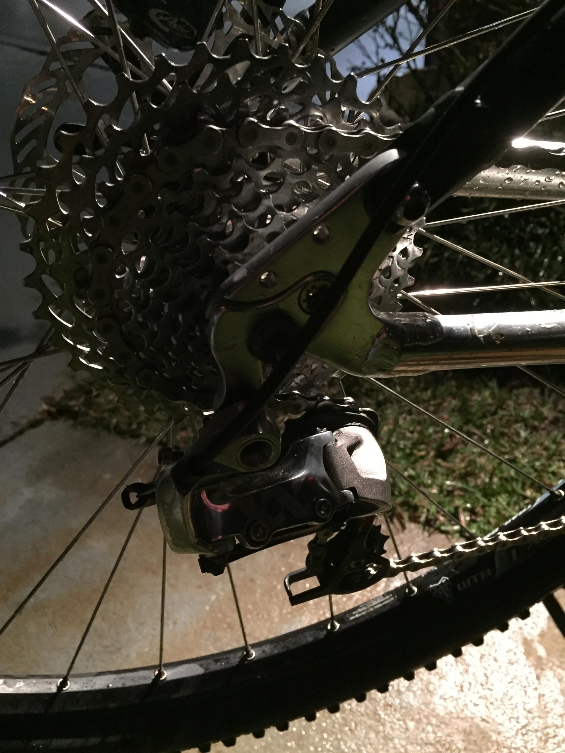 Voodoo Cycles Canzo 29er F/S: Close-up image of a bicycle's rear cassette and derailleur, showing the detailed metal components and chain, with a blurred background suggesting nighttime conditions. The surface beneath the bike is wet, reflecting light.