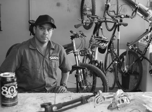 Specialized Stumpjumper FSR: A man wearing a work shirt with the logo "SURLY" sits at a table in a bike shop. Behind him, several bicycles are displayed, and a can of beer is resting on the table. The image is in black and white, highlighting the man's focused expression amidst the tools and bike parts around him.