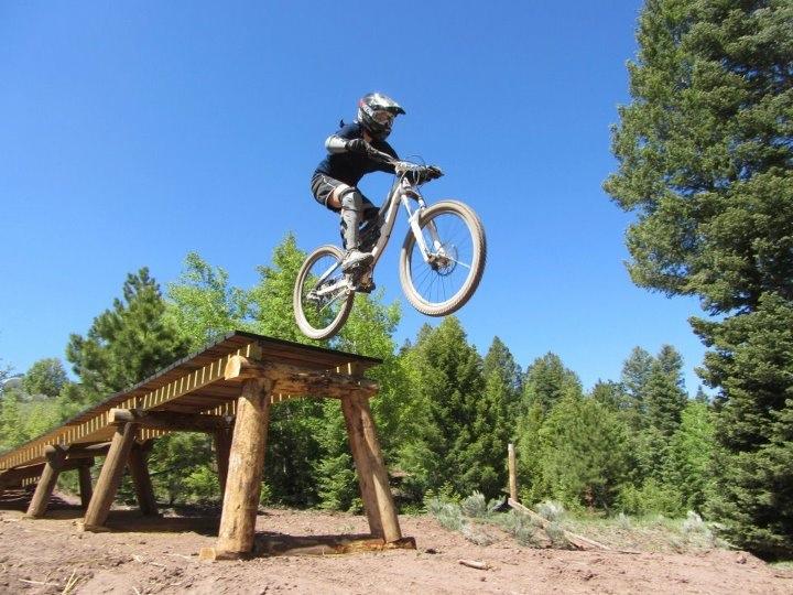 A mountain biker dressed in protective gear performs a jump off a wooden ramp, surrounded by dense greenery under a clear blue sky. Angel Fire Bike Park mountain bike trail.