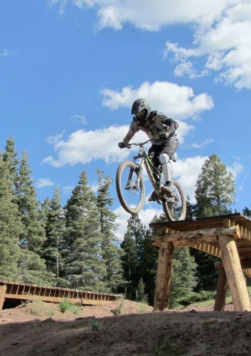 A mountain biker in protective gear jumps off a wooden ramp, soaring over a dirt path with trees and a blue sky in the background. Angel Fire Bike Park mountain bike trail.