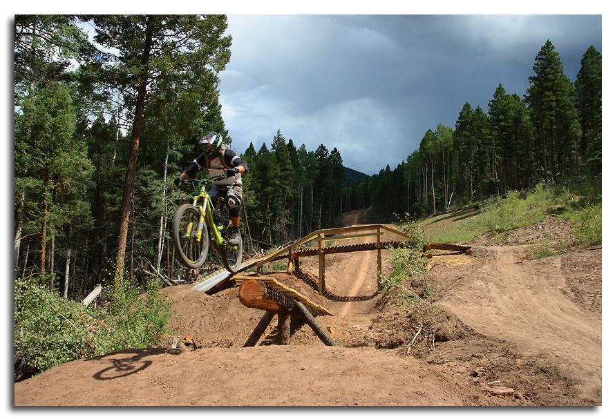 A mountain biker performing a jump over a wooden ramp on a dirt trail in a forested area, with tall trees and cloudy skies in the background. Angel Fire Bike Park mountain bike trail.