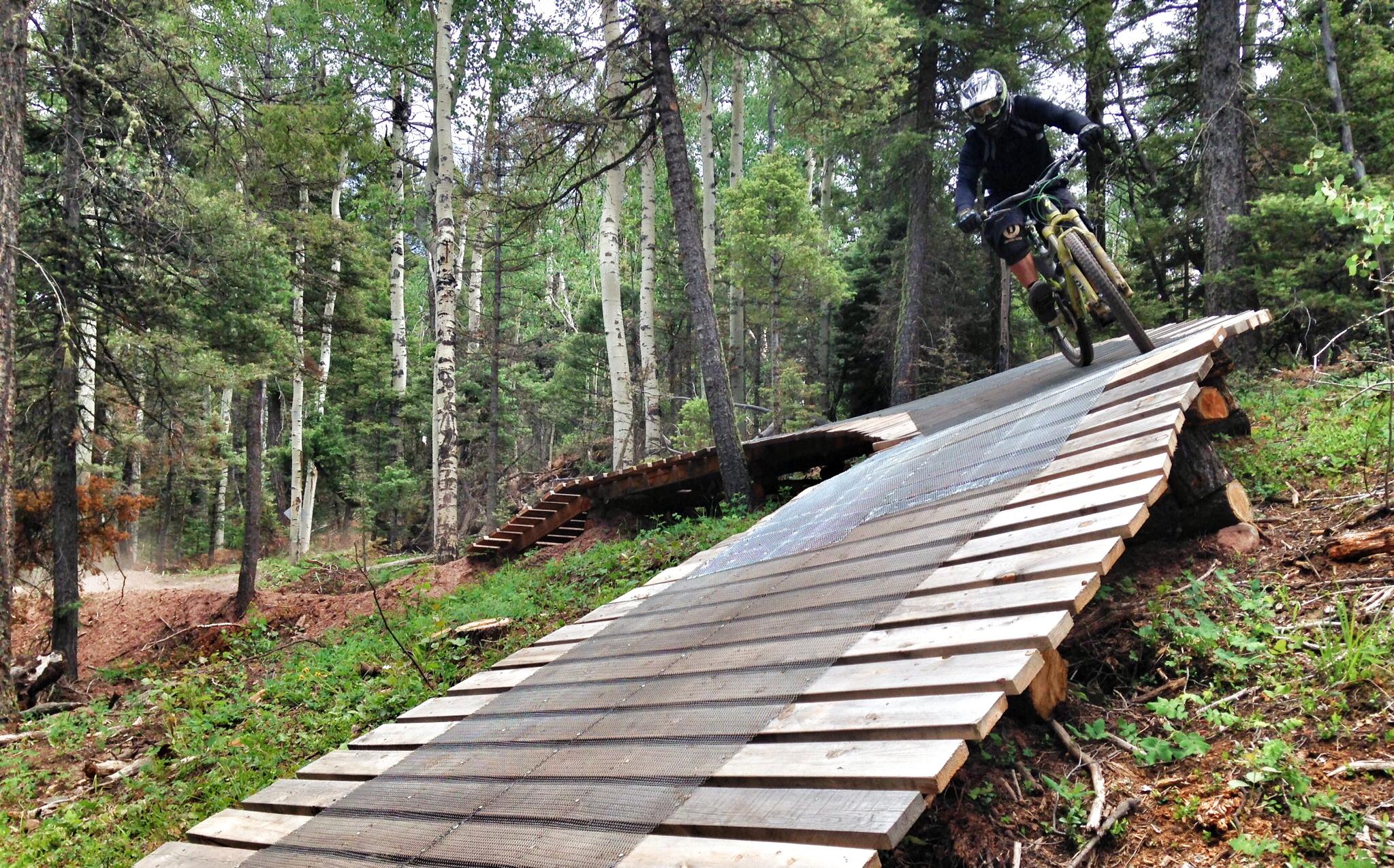 A mountain biker riding down a wooden ramp in a forested area, surrounded by tall trees and greenery. The ramp is designed for biking, featuring a sturdy wooden surface with a mesh section. Dust is visible in the air, indicating speed and movement. Angel Fire Bike Park mountain bike trail.